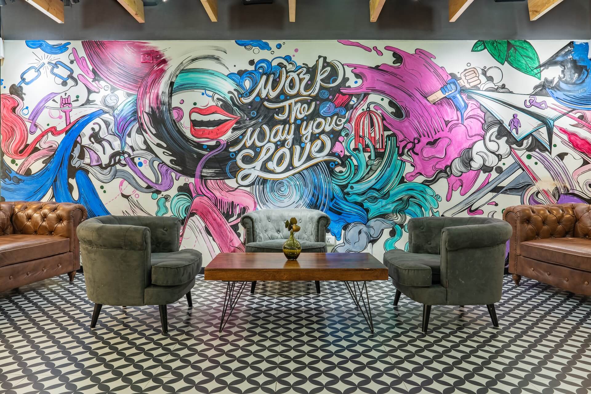 Amplo lounge de coworking com sofa de couro marrom nas laterais duas poltronas verdes e mesa de centro de madeira. Parede grafitada apresenta desenhos fluidos em azul rosa roxo com lettering Work the way you love