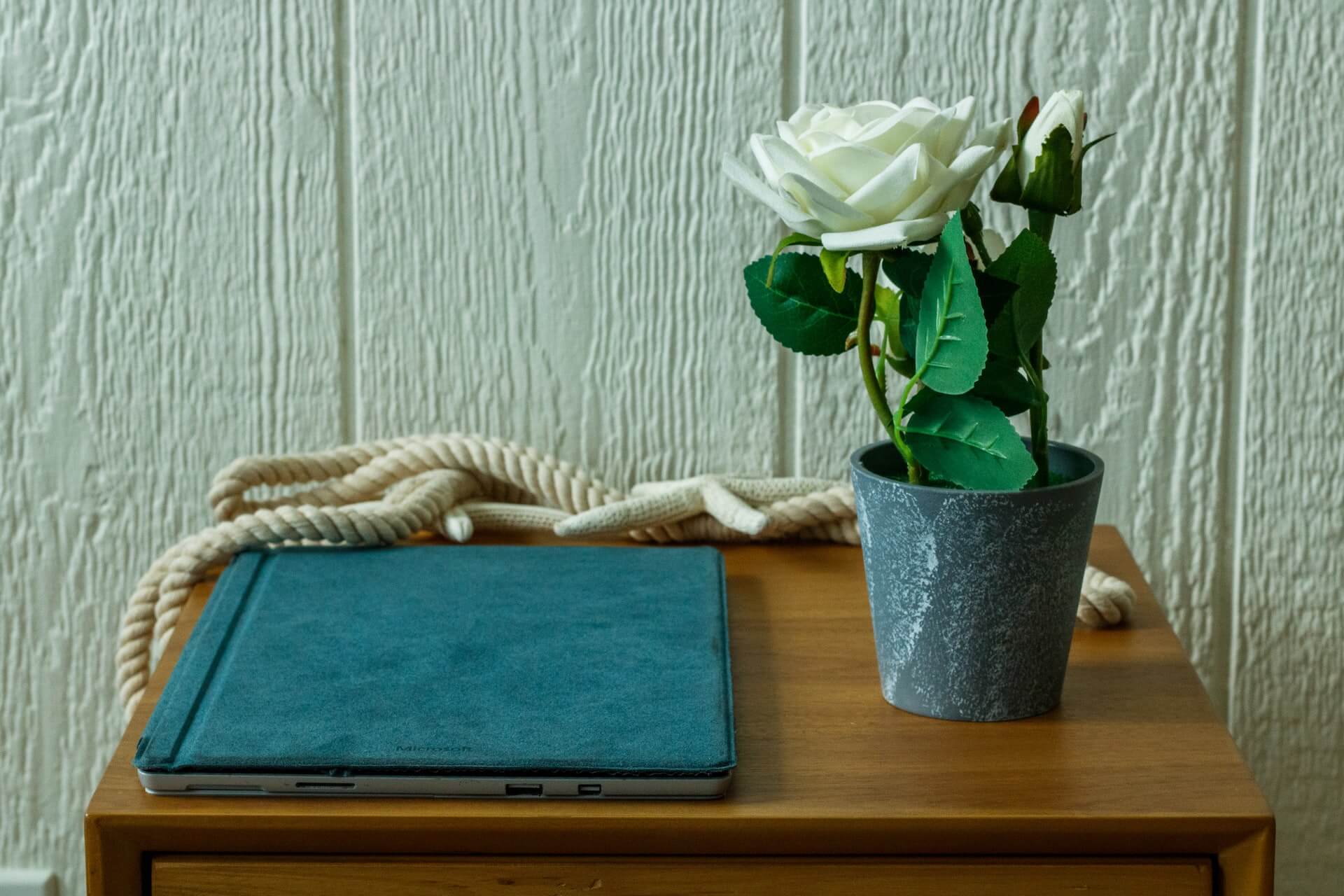 Mesa de madeira com portatil fechado com capa azul, corda enrolada e vaso cinzento com rosa branca artificial