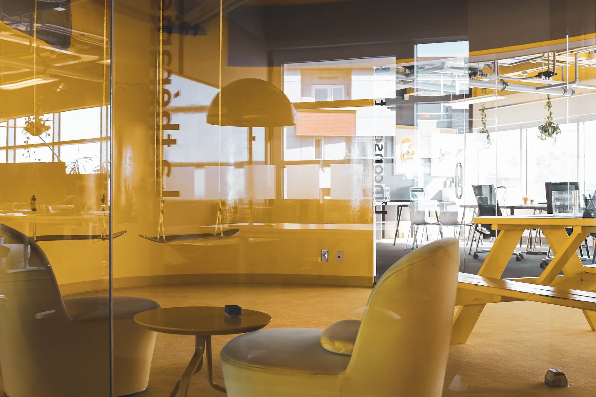 Vista de área de co-living/coworking através de vidro amarelo com cadeirão, mesa de piquenique suspensa, candeeiro pendente e mesas de trabalho ao fundo