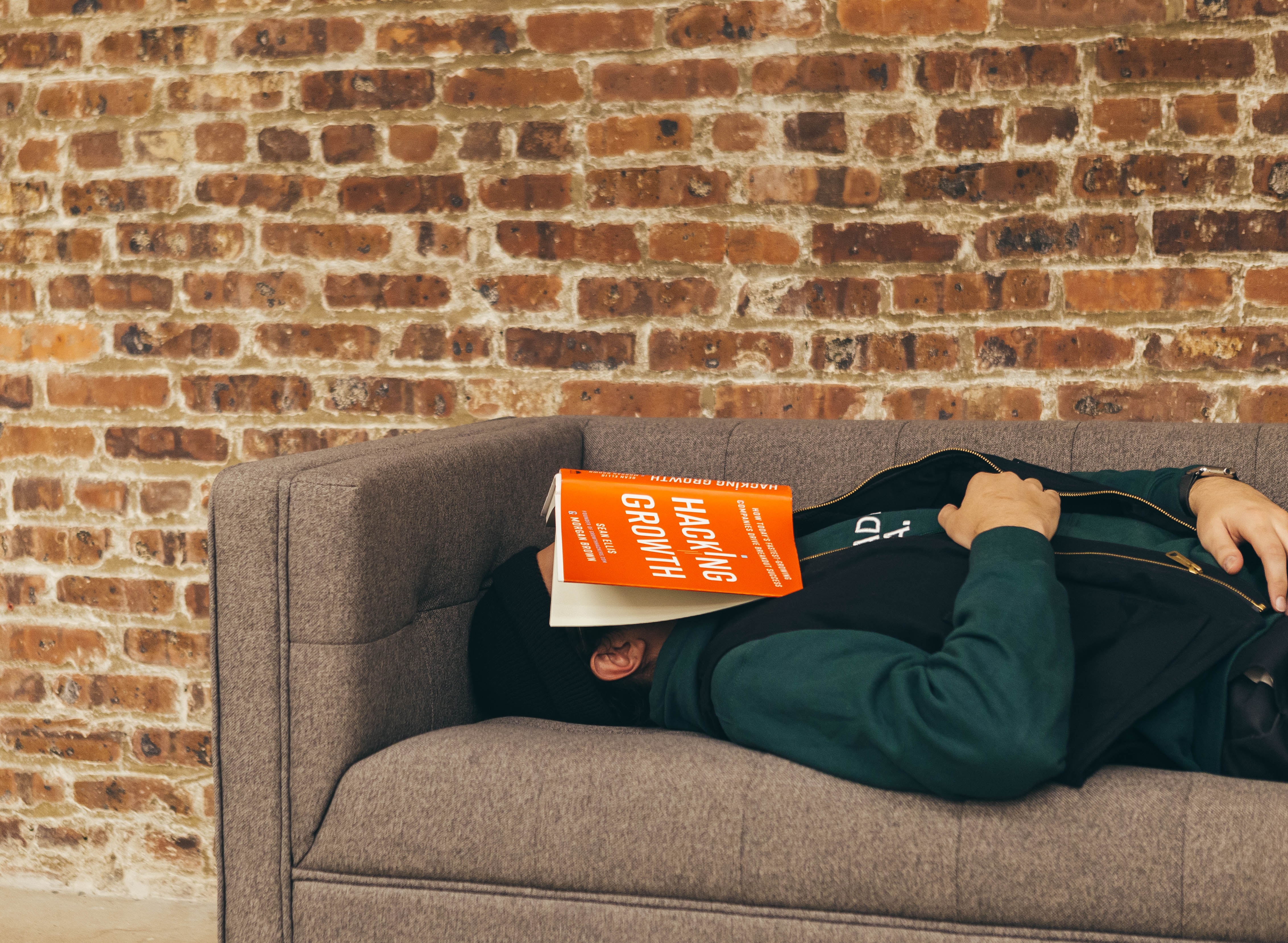 Pessoa vestindo casaco verde e gorro preto deitada num sofá cinzento com o livro de capa laranja Hacking Growth sobre o rosto e parede de tijolos ao fundo