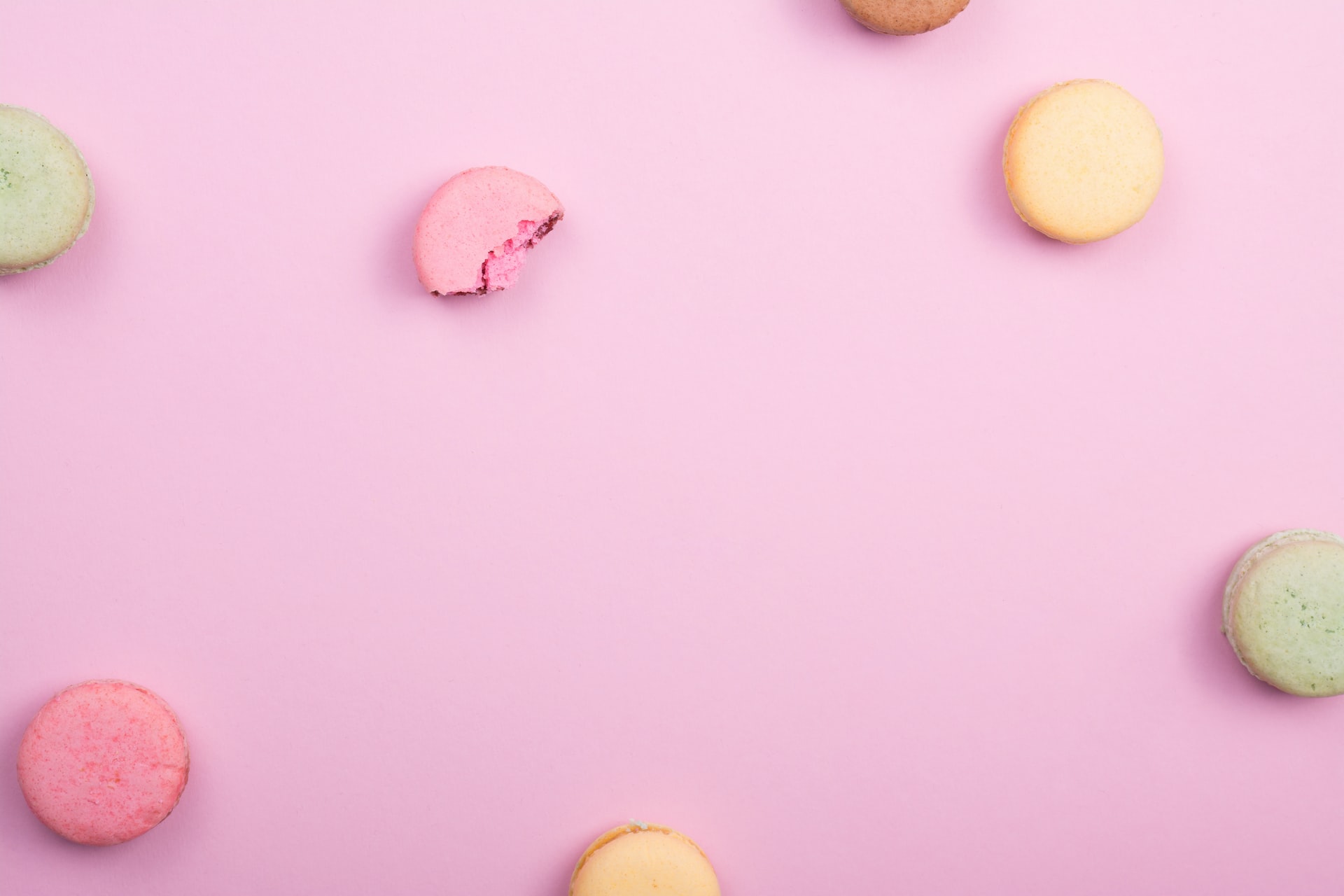 Vista aérea de macarons coloridos (rosa, amarelo, verde), um deles parcialmente comido, dispostos em torno de espaço vazio sobre fundo rosa
