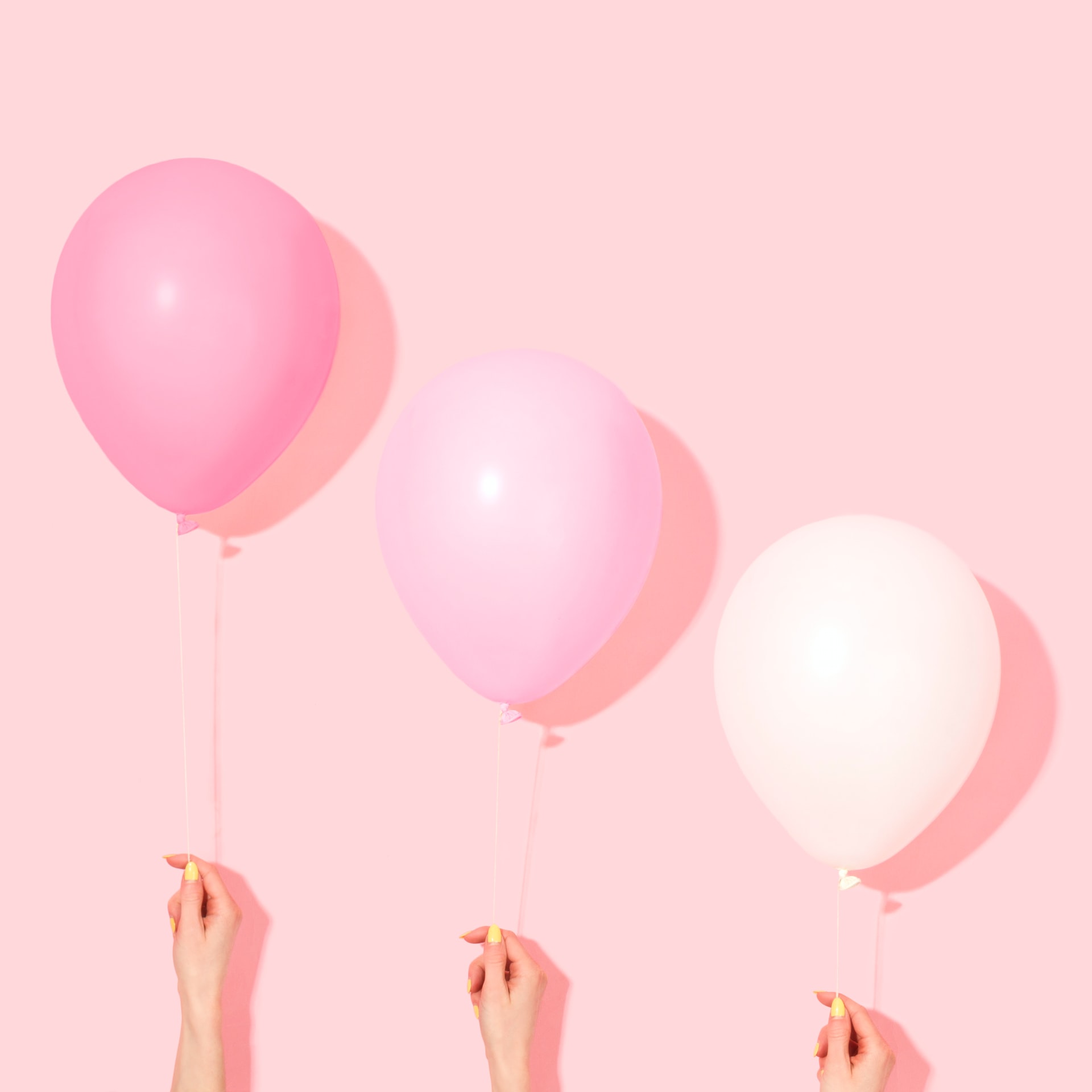 Três balões nas cores rosa-escuro rosa-claro e branco suspensos por mãos com unhas amarelas sobre fundo rosa pastel