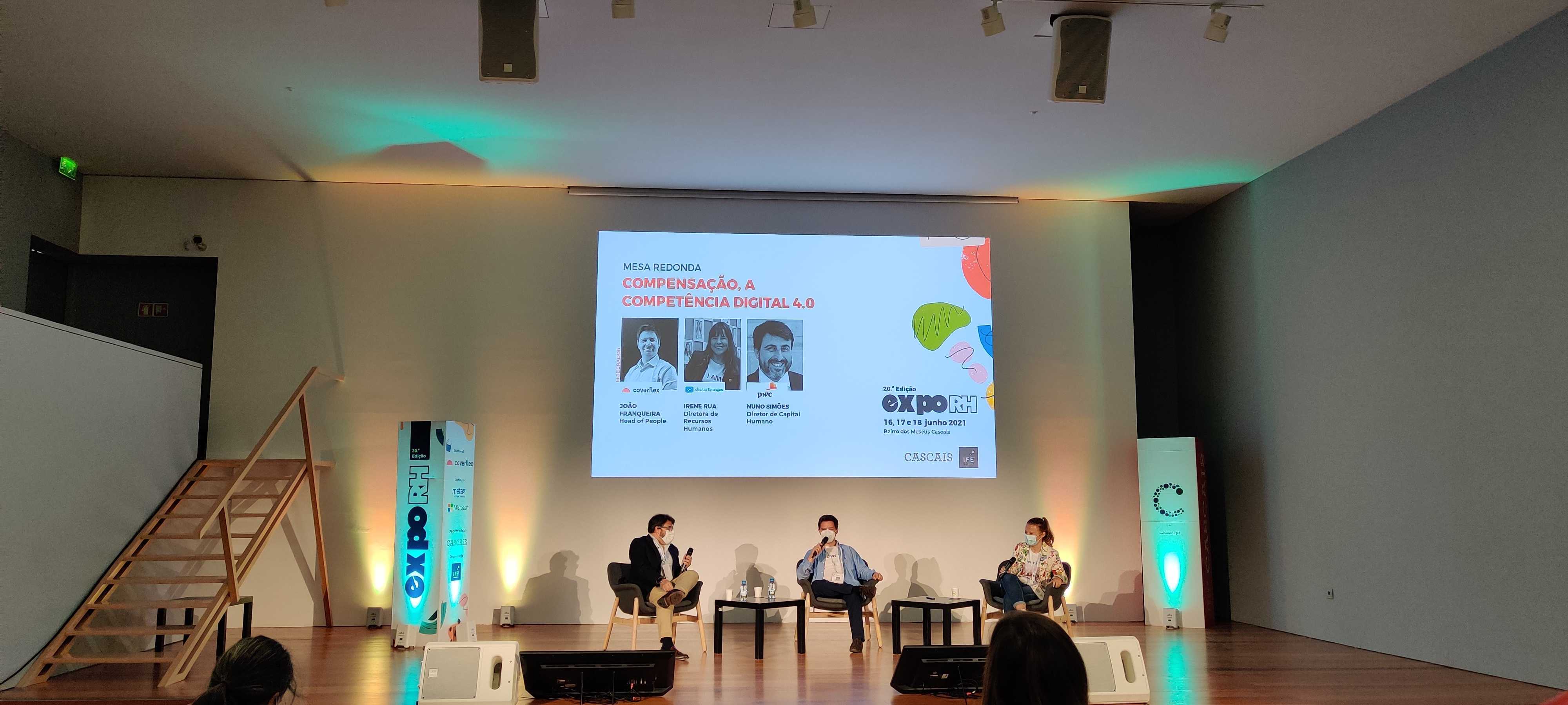 Três participantes usam máscara sentados em cadeiras no palco do Expo RH em sala iluminada com colunas laterais e ecrã central com título Mesa Redonda compensação a competência digital 4 ponto 0 e fotos de oradores