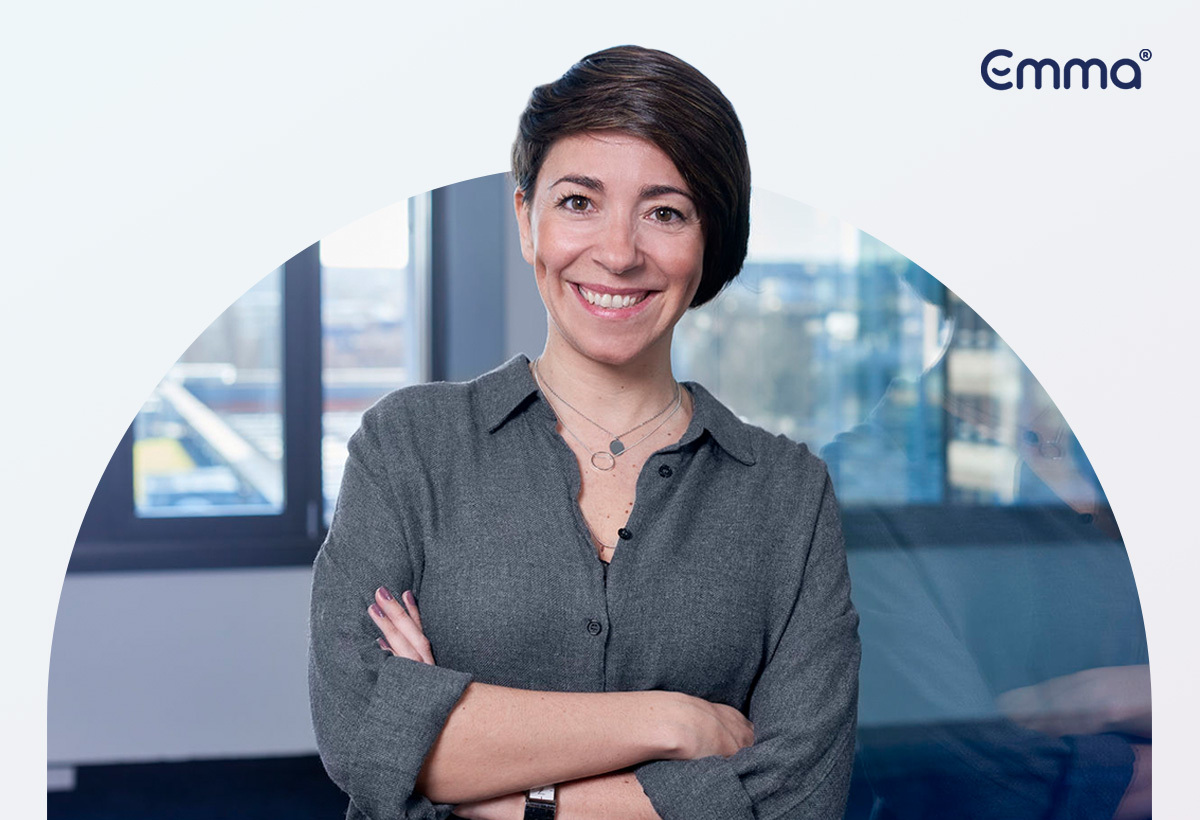 Mulher sorridente de braços cruzados em escritório com logo Emma no canto superior direito