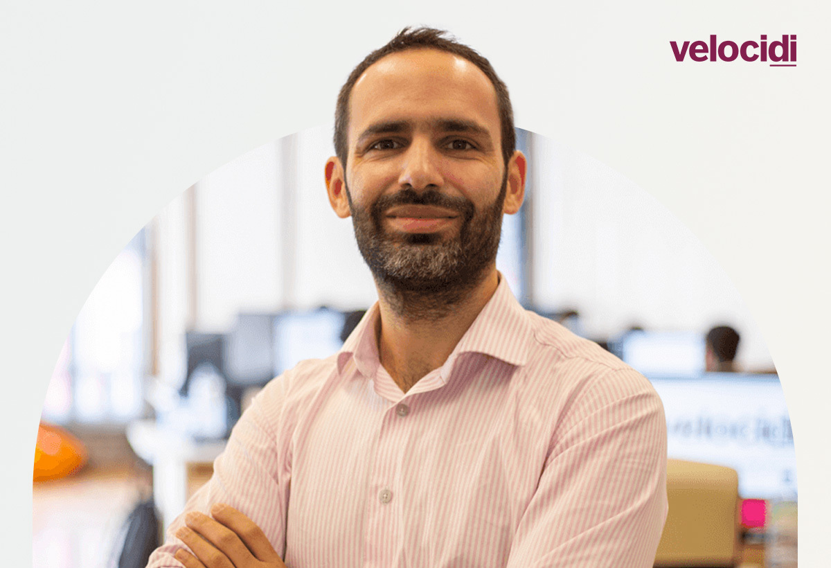 Paulo Cunha, CEO da Velocidi, de braços cruzados e sorridente num escritório com computadores ao fundo e logotipo da Velocidi no canto superior direito