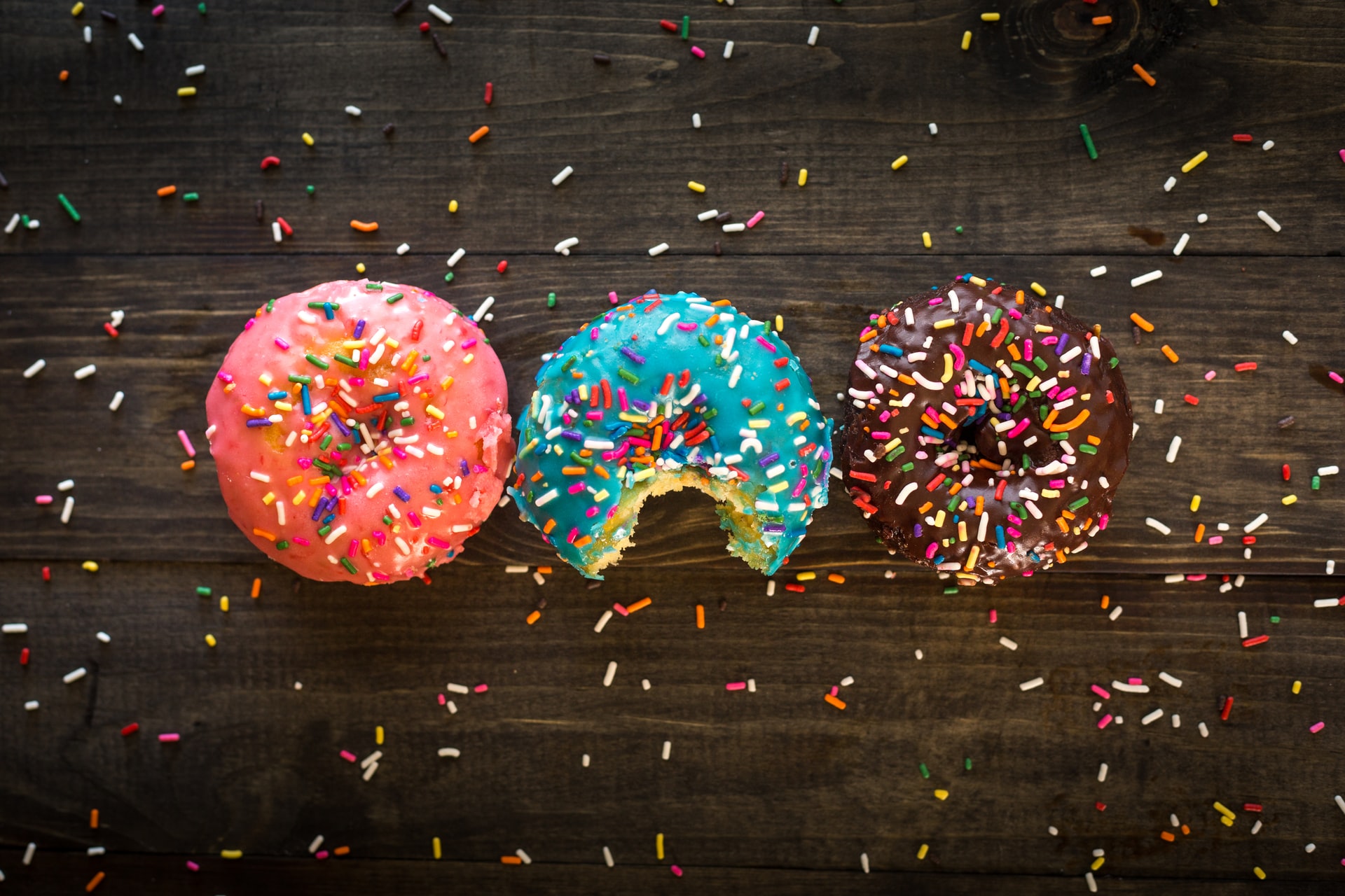 Três donuts alinhados sobre tábua de madeira: um com cobertura rosa, outro azul com um pedaço mordido e um de chocolate, todos polvilhados com granulados coloridos e migalhas espalhadas ao redor