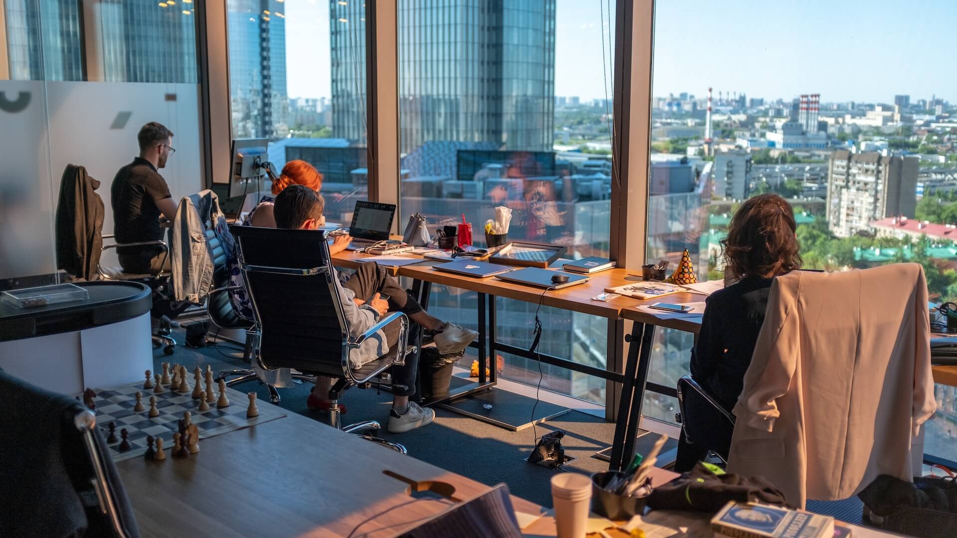 Open office space with employees working at desks, laptops and books on the tables, large windows offering a panoramic urban view, and a chessboard in the foreground