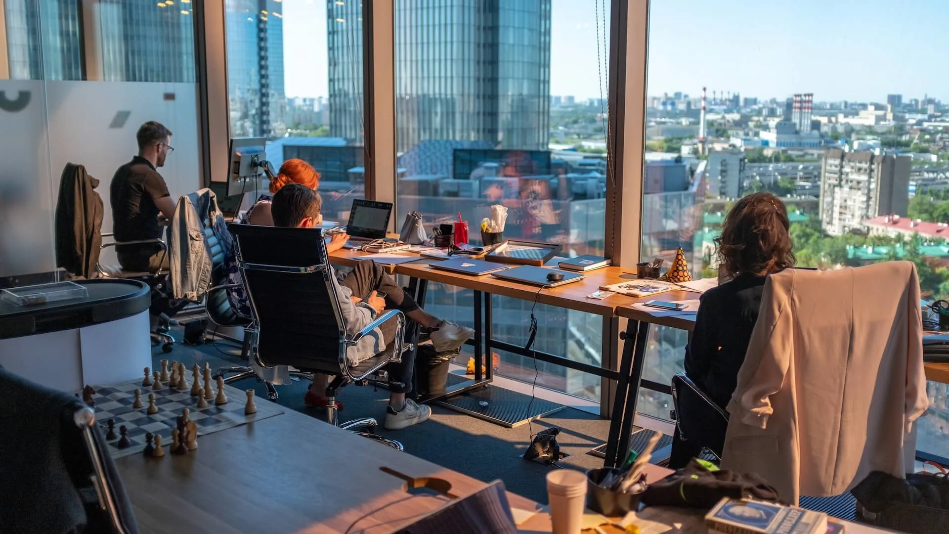 Open office space with employees working at desks, laptops and books on the tables, large windows offering a panoramic urban view, and a chessboard in the foreground