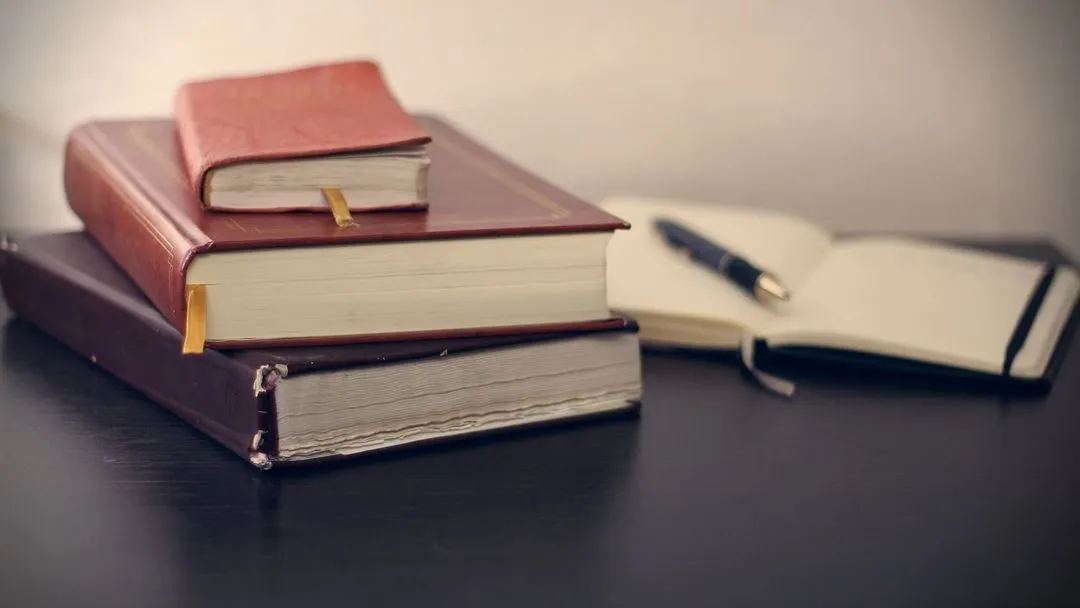 A stack of three hardcover books with ribbons on a dark desk, next to an open notebook and a pen, suggesting a study or work environment