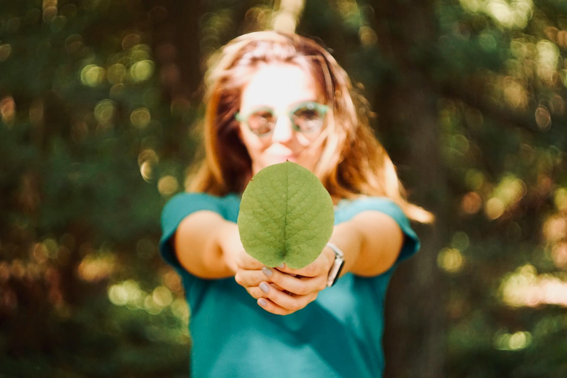 Mulher com óculos de sol segurando uma folha verde em destaque, com fundo de floresta desfocado ao sol