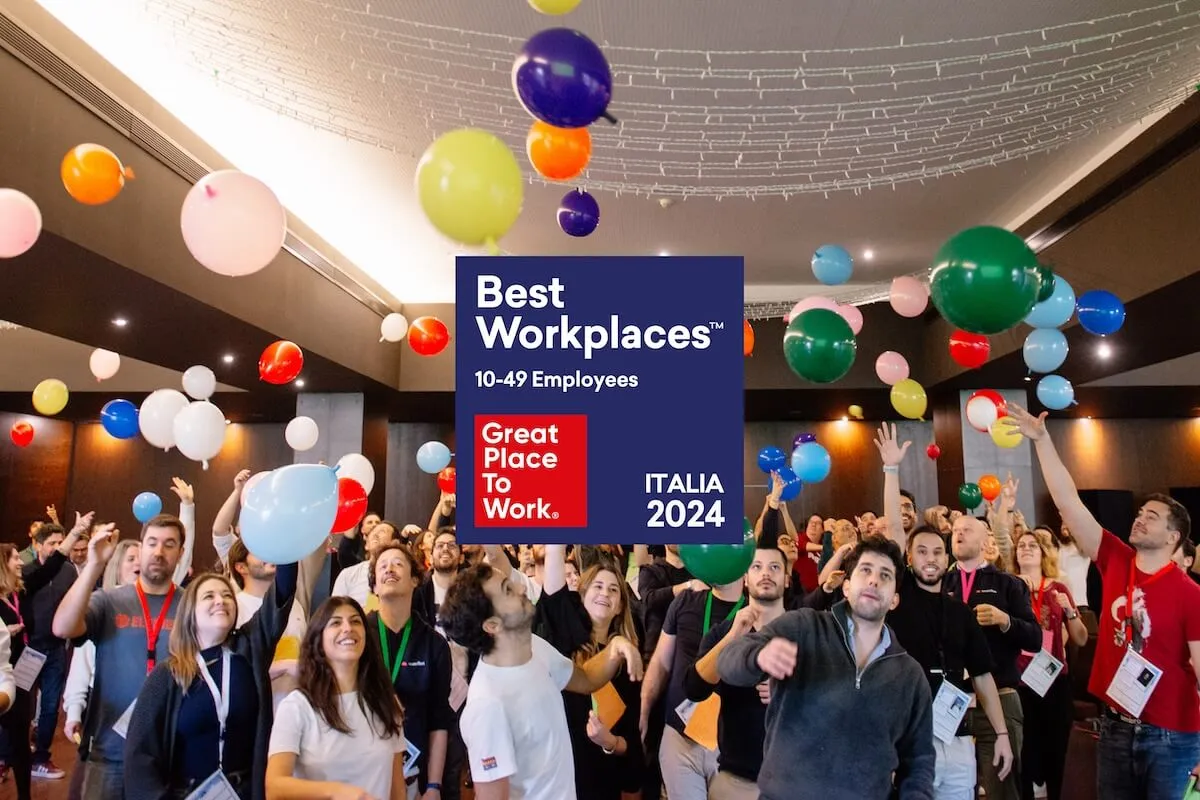 Várias pessoas reunidas num salão celebrando com balões coloridos no ar. No centro da imagem, há um destaque visual de reconhecimento Best Workplaces 10-49 Employees Great Place to Work Italia 2024.