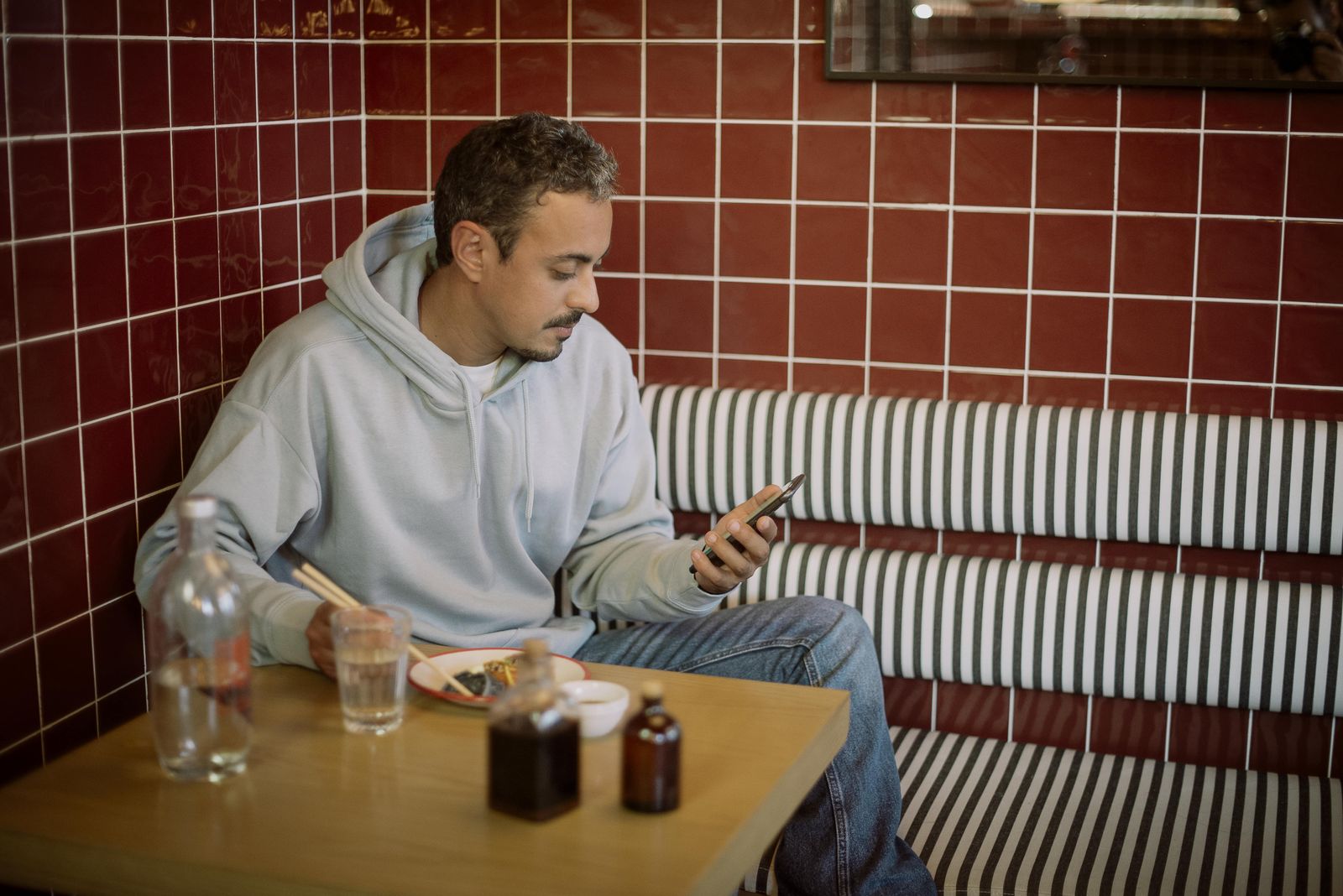 Casual diner wearing a grey hoodie and jeans sits in a red tiled booth at a wooden table using chopsticks to eat noodles while looking at his smartphone with bottles and a glass of water on the table