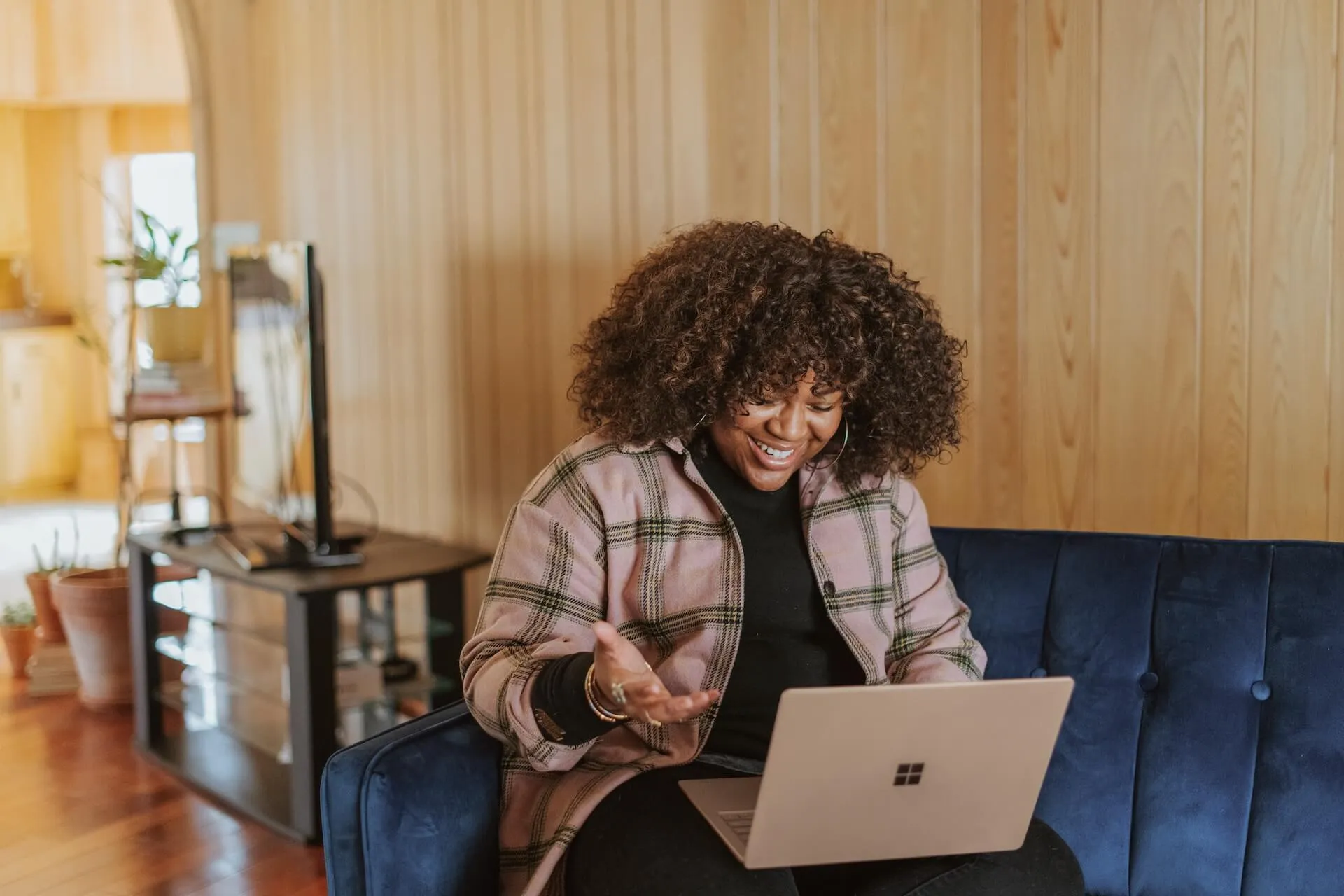 Black woman wearing a plaid jacket sits on a navy sofa at home, smiling and gesturing while working on a laptop, with wood paneled walls, plants and a TV stand in the background