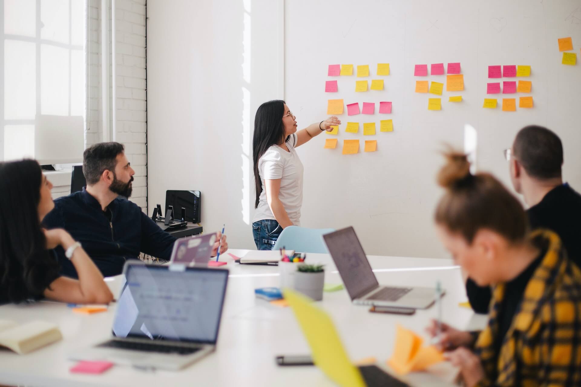 a diverse team sits around a conference table covered in laptops and notebooks while a woman stands at a white wall arranging pink and yellow sticky notes during a collaborative brainstorming session in a bright office