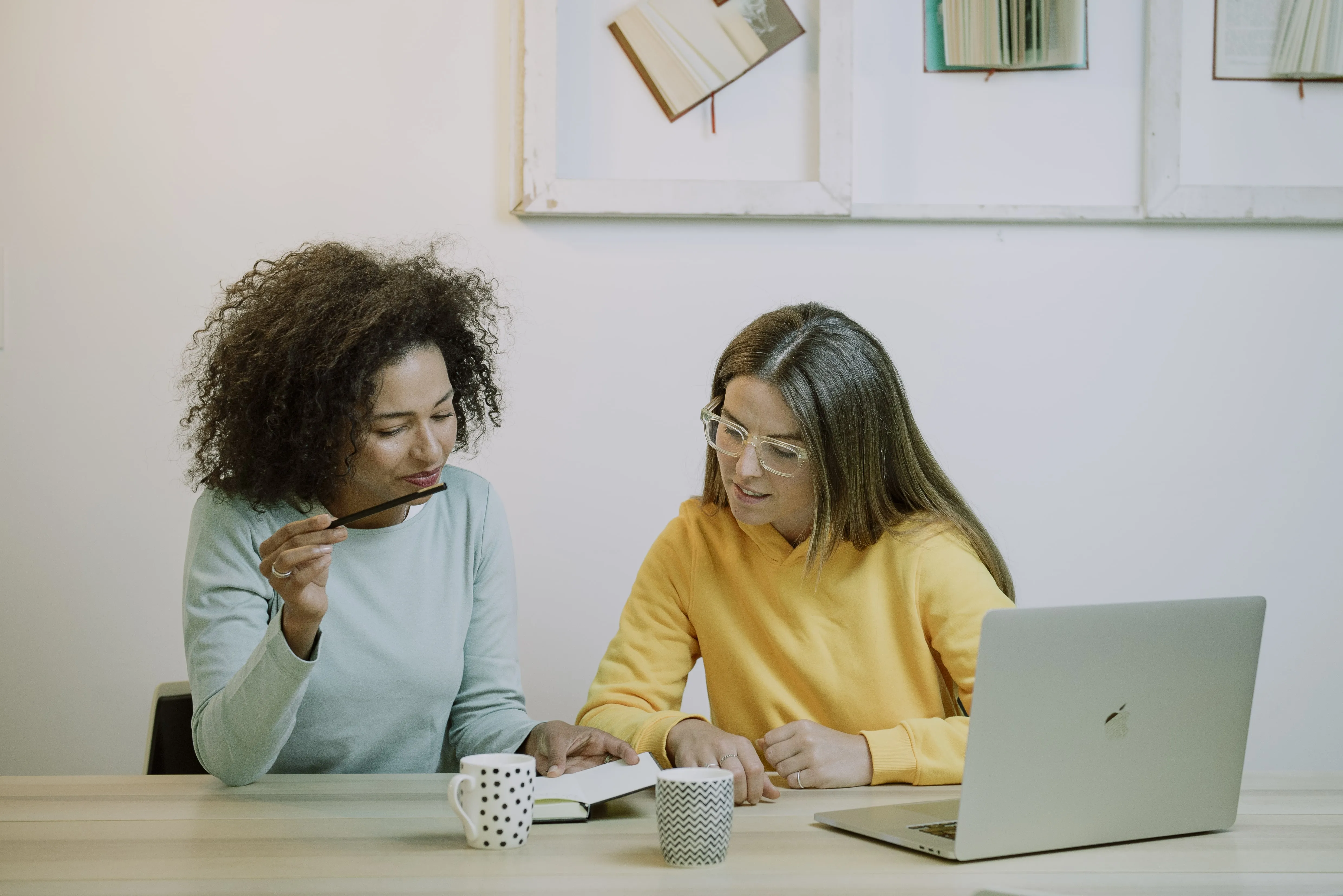 Two women in a bright workspace sitting at a wooden table with coffee mugs and a laptop while discussing and pointing at a small open notebook