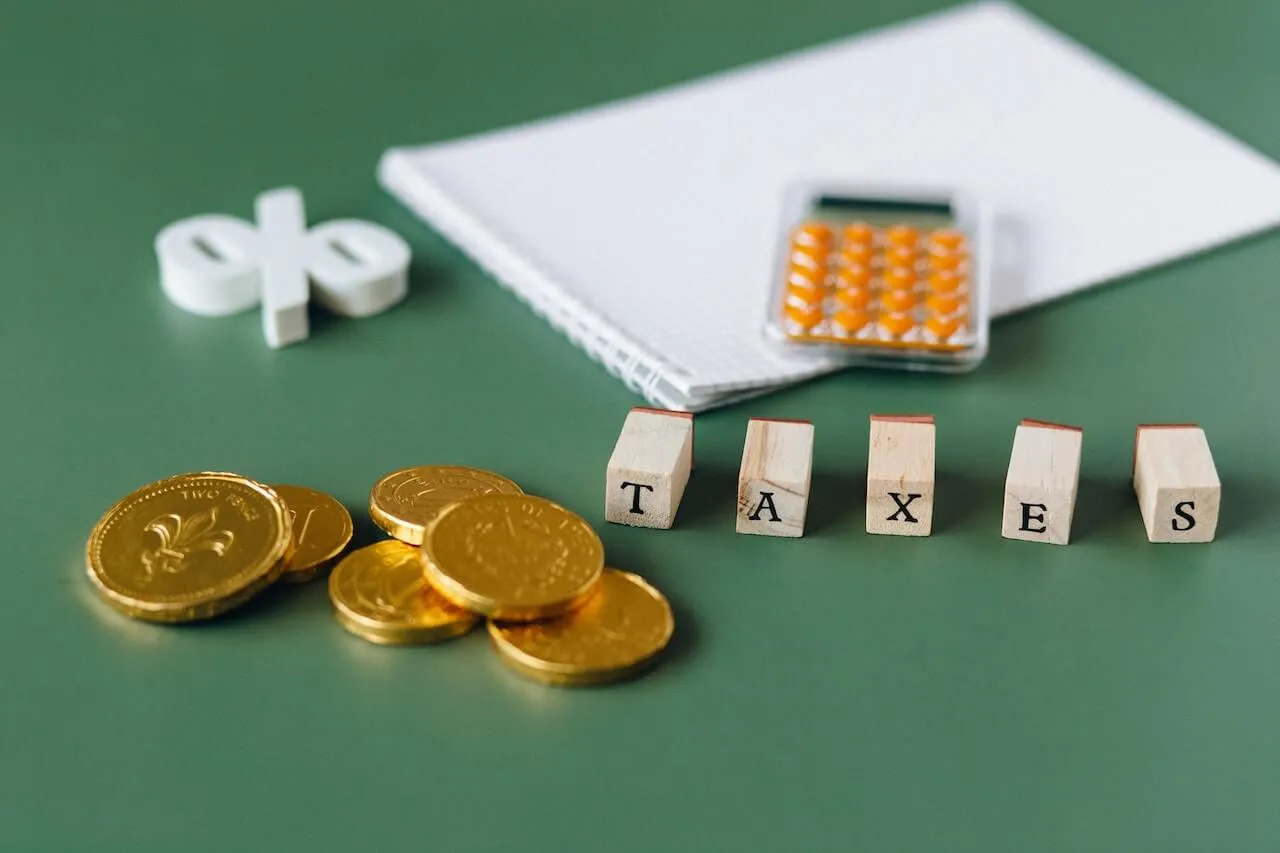 On a green desk wooden letter blocks spelling TAXES surrounded by a stack of gold coins a white percent sign a grid notebook and an orange calculator representing tax benefits
