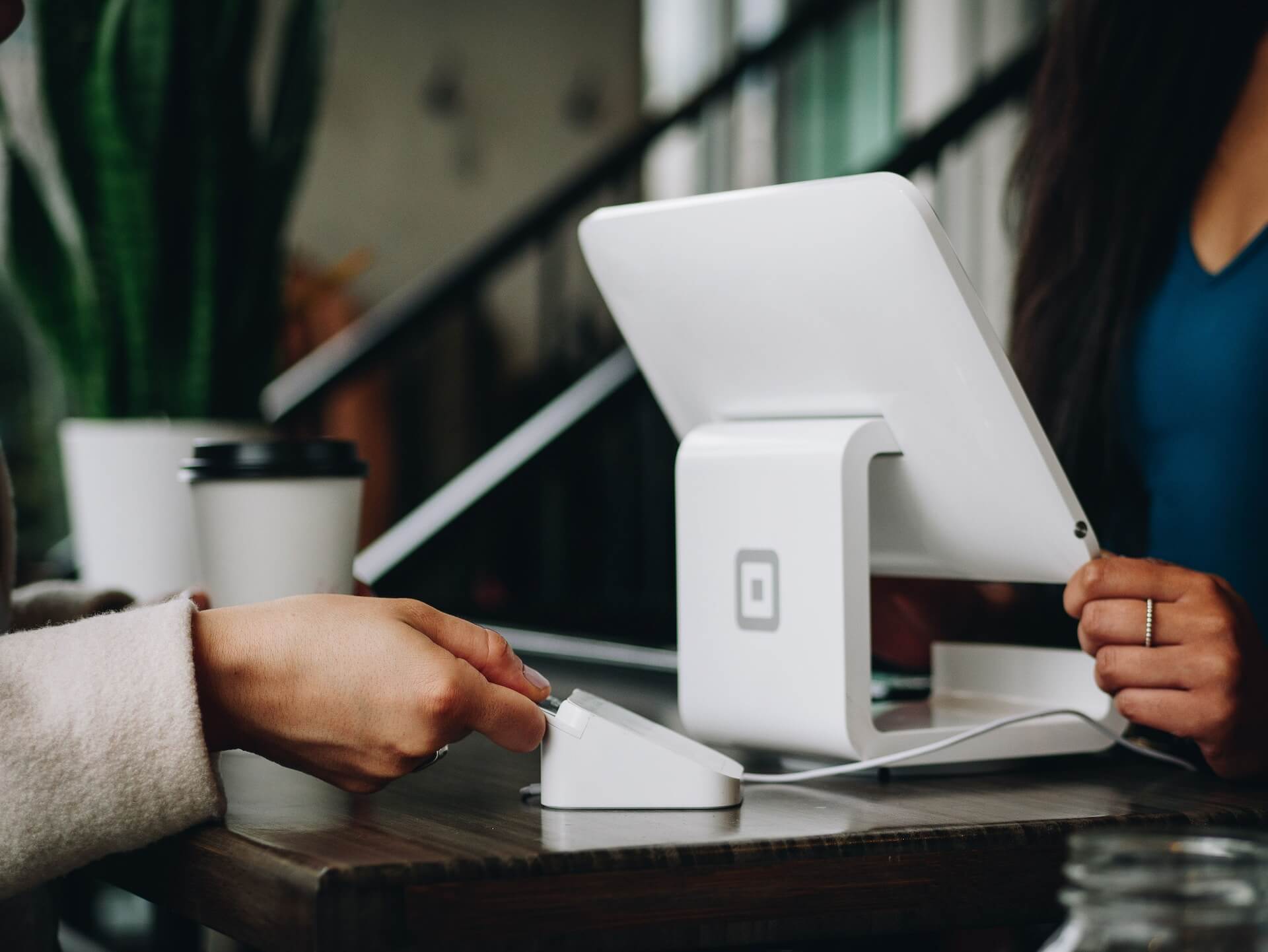 Customer taps payment card on a white card reader attached to a white touchscreen POS on a wooden counter with coffee cup and blurred background