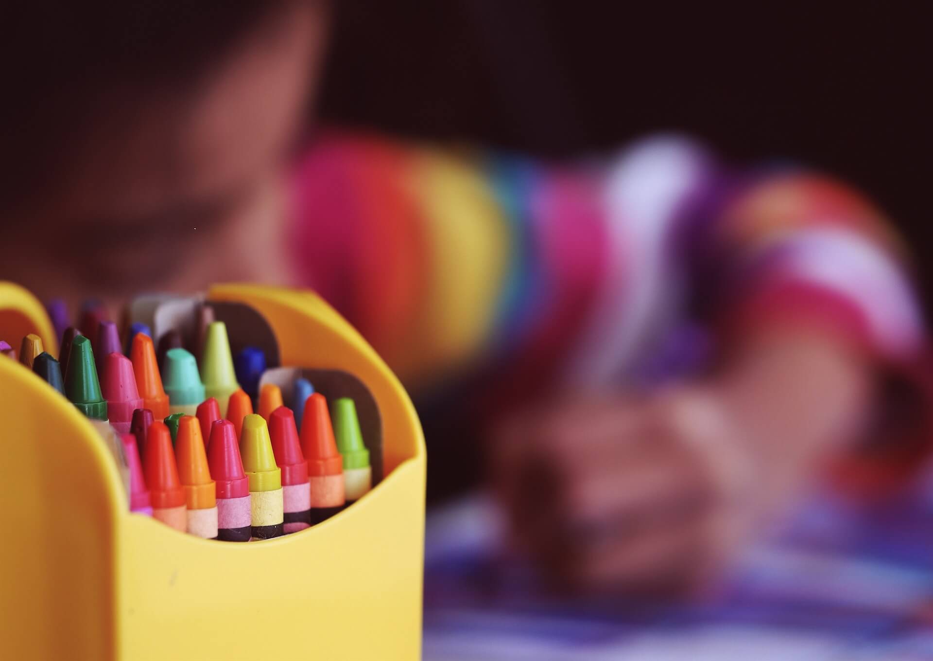 Yellow box filled with assorted crayons in sharp focus and a child in a rainbow striped sweater drawing in the blurred background