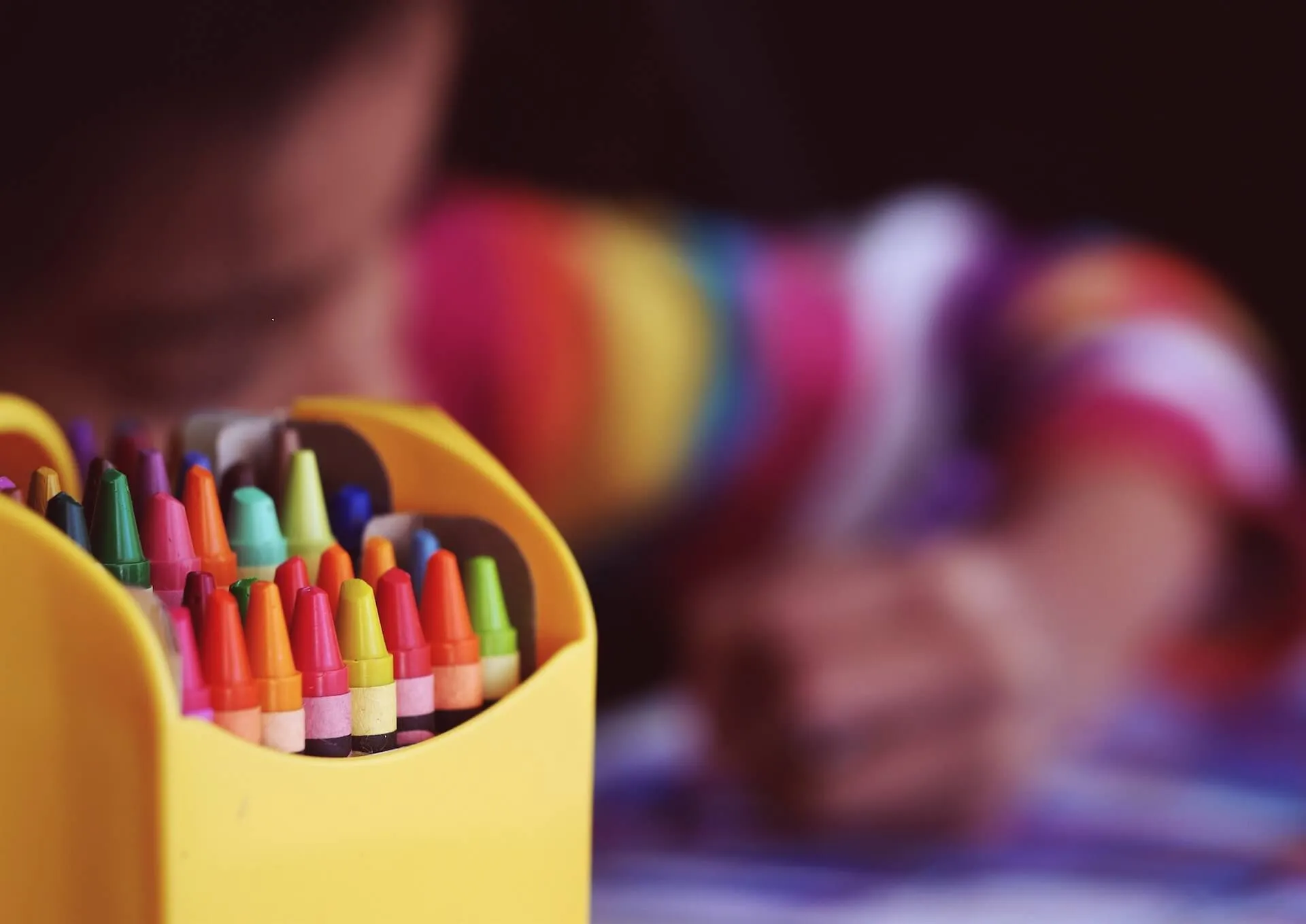 Yellow box filled with assorted crayons in sharp focus and a child in a rainbow striped sweater drawing in the blurred background