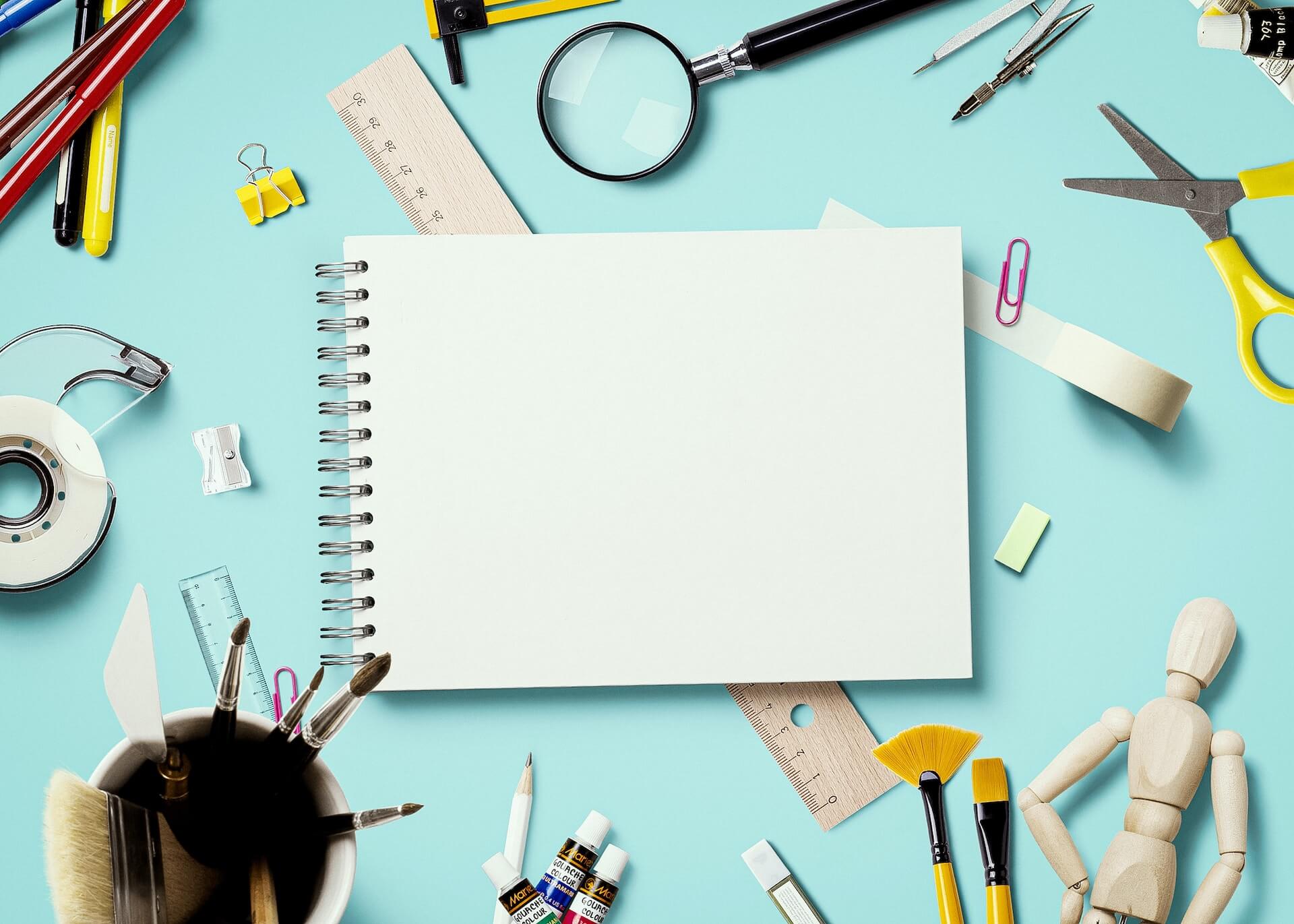 Top view of a white spiral sketchbook in the center with pencils markers rulers paint brushes tubes of paint scissors tape magnifying glass a wooden mannequin and paper clips arranged around it on cyan surface