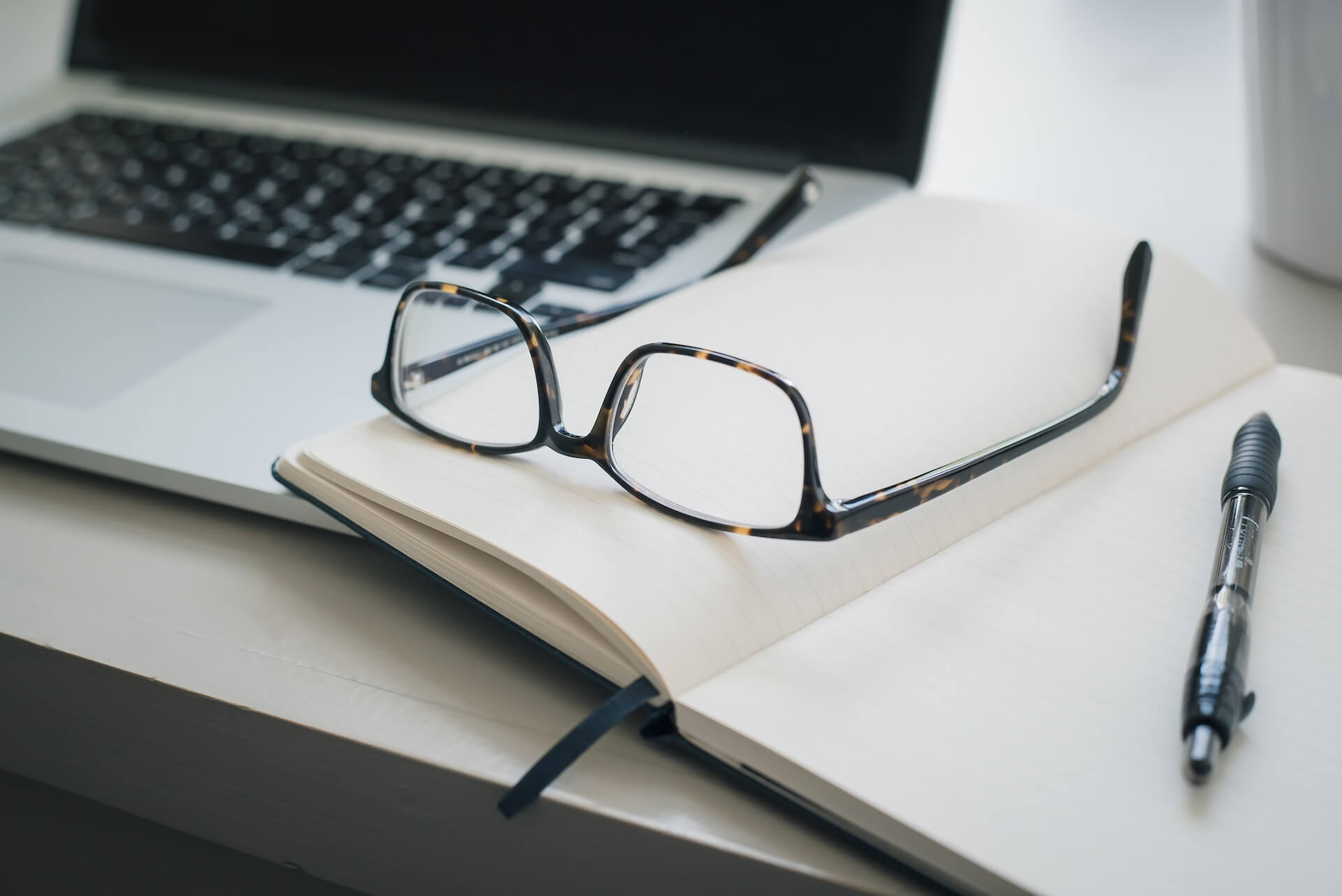 close-up of a workspace showing tortoiseshell eyeglasses resting on blank pages of an open notebook next to a laptop keyboard and a black pen