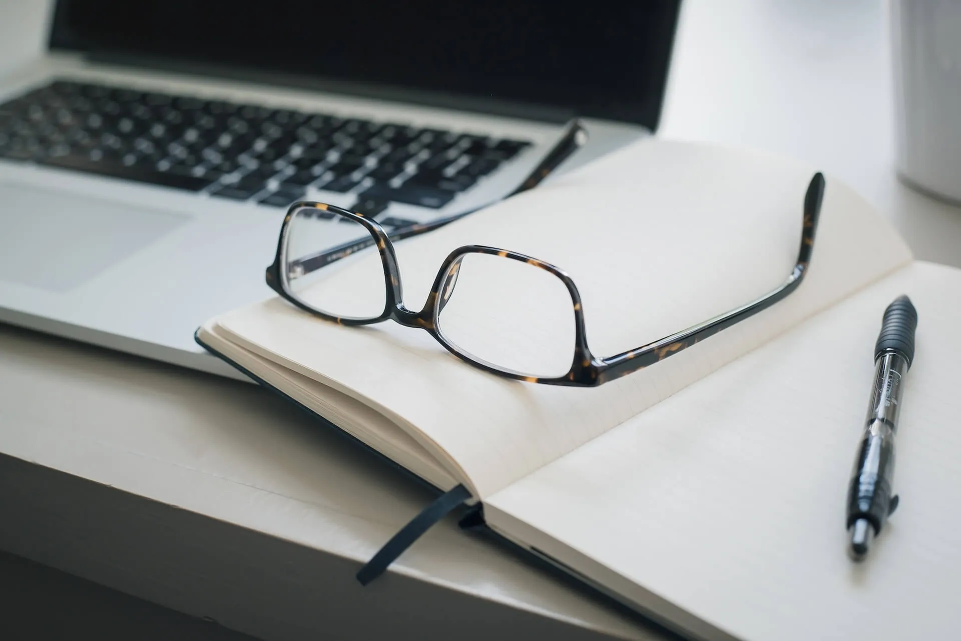 close-up of a workspace showing tortoiseshell eyeglasses resting on blank pages of an open notebook next to a laptop keyboard and a black pen