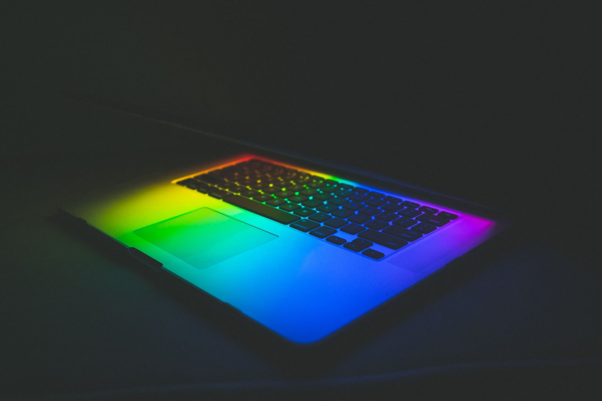 overhead view of a laptop keyboard and trackpad illuminated by a rainbow spectrum of light in a dark setting