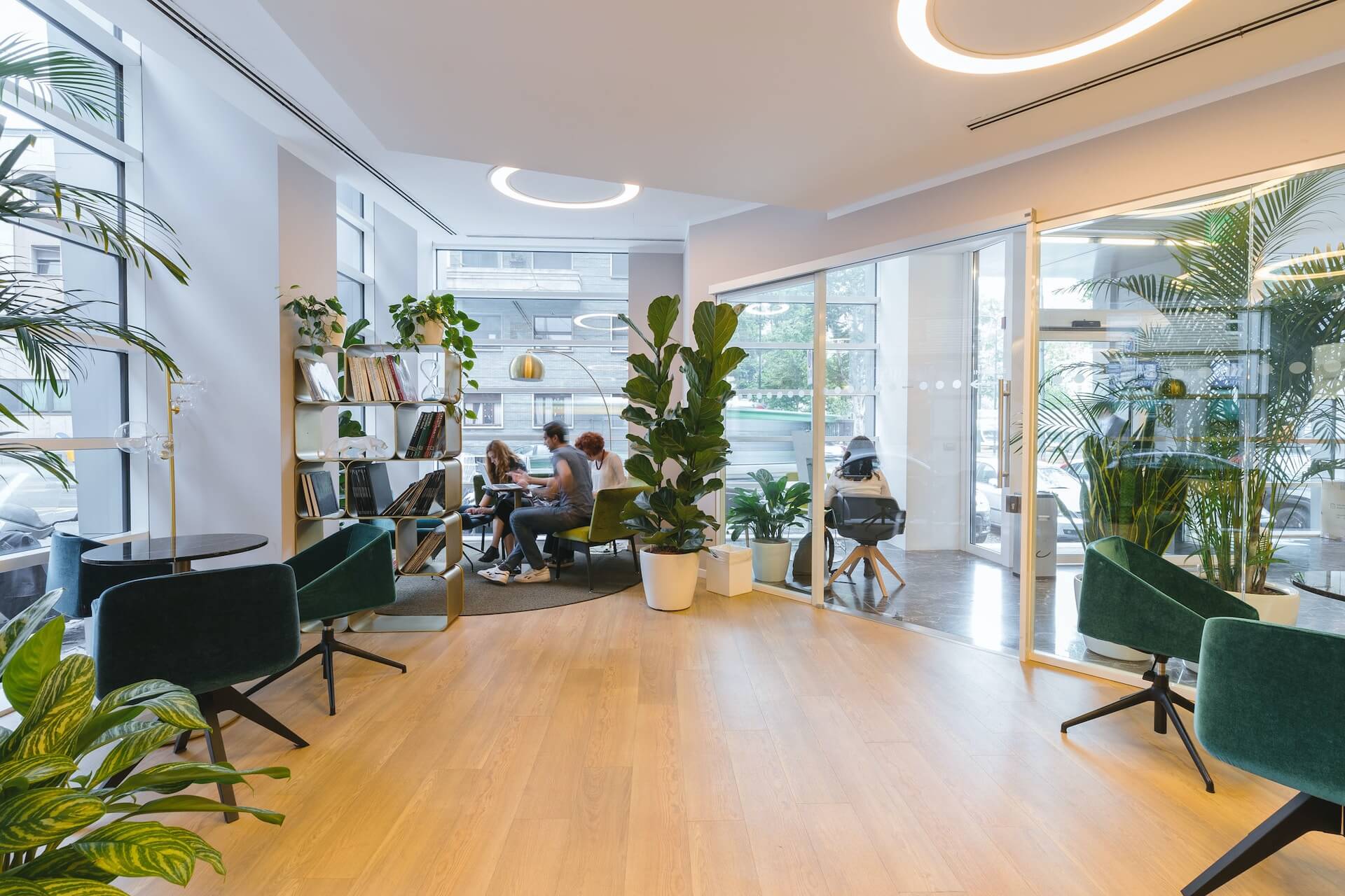 Open office reception area with wooden floor green lounge chairs a bookshelf room divider potted plants a glass meeting room and people collaborating at a table by large windows