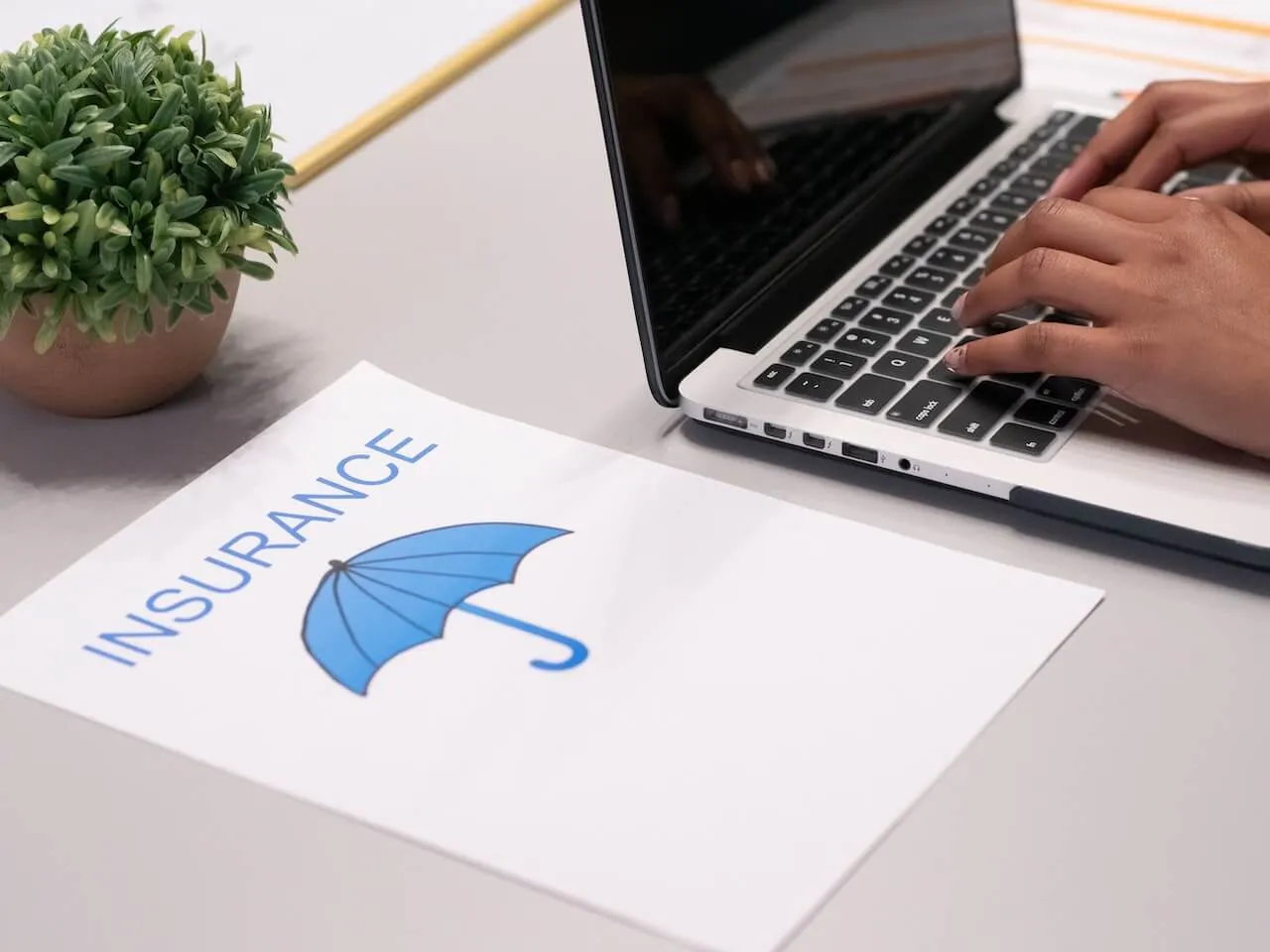 close up of person typing on a laptop beside an insurance document featuring a blue umbrella icon and a small plant on a desk
