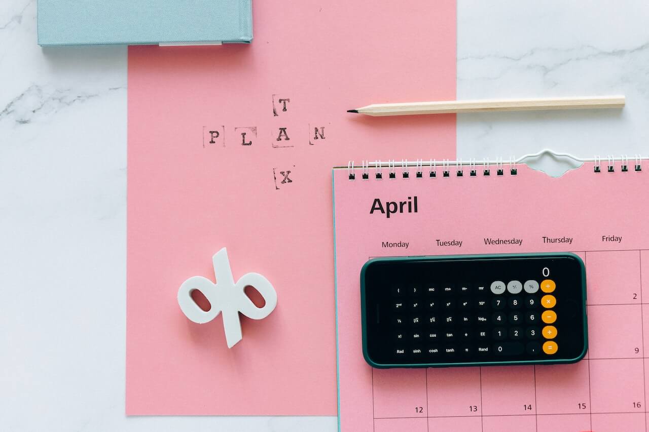 Overhead view of pastel pink paper and an April calendar page topped by a smartphone calculator, light wood pencil, white 3D percent sign and black stamped letters spelling TAX PLAN