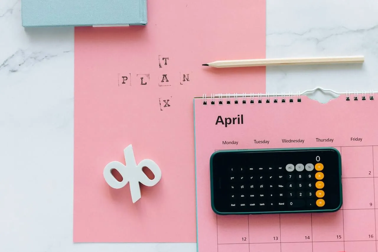 Overhead view of pastel pink paper and an April calendar page topped by a smartphone calculator, light wood pencil, white 3D percent sign and black stamped letters spelling TAX PLAN