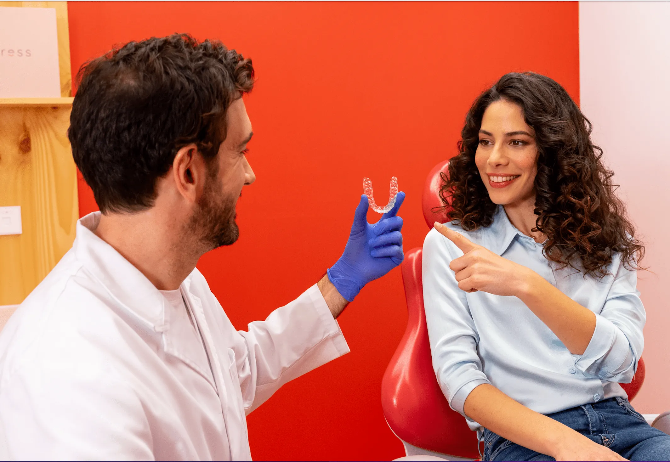 Male orthodontist in white coat and blue gloves holds transparent aligner as smiling female patient points at it while seated in a red dental chair