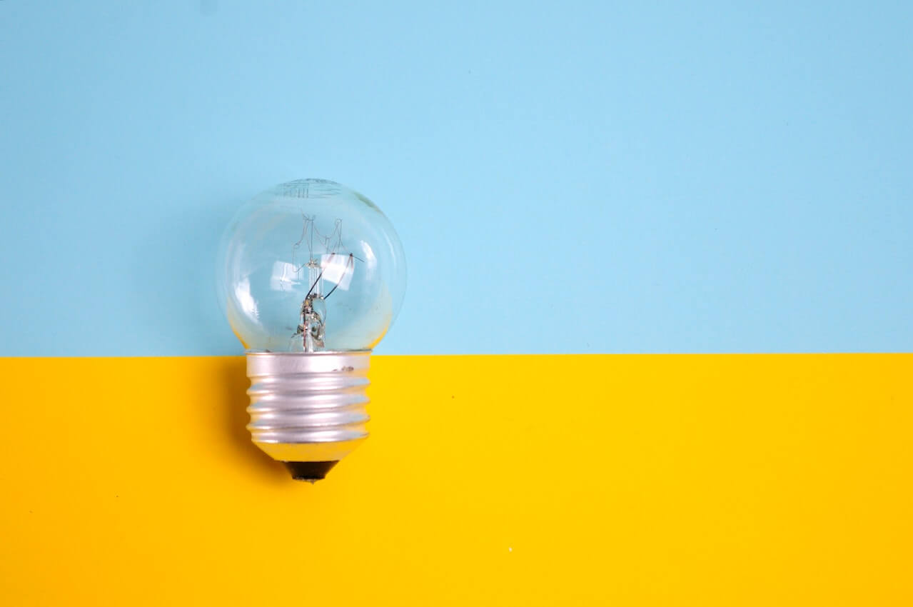 single clear incandescent light bulb resting on a horizontal split background with light blue on top and bright yellow on bottom