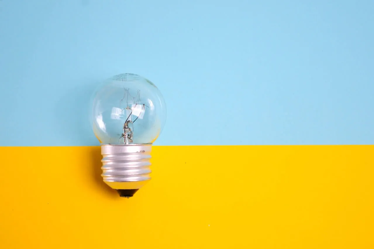 single clear incandescent light bulb resting on a horizontal split background with light blue on top and bright yellow on bottom