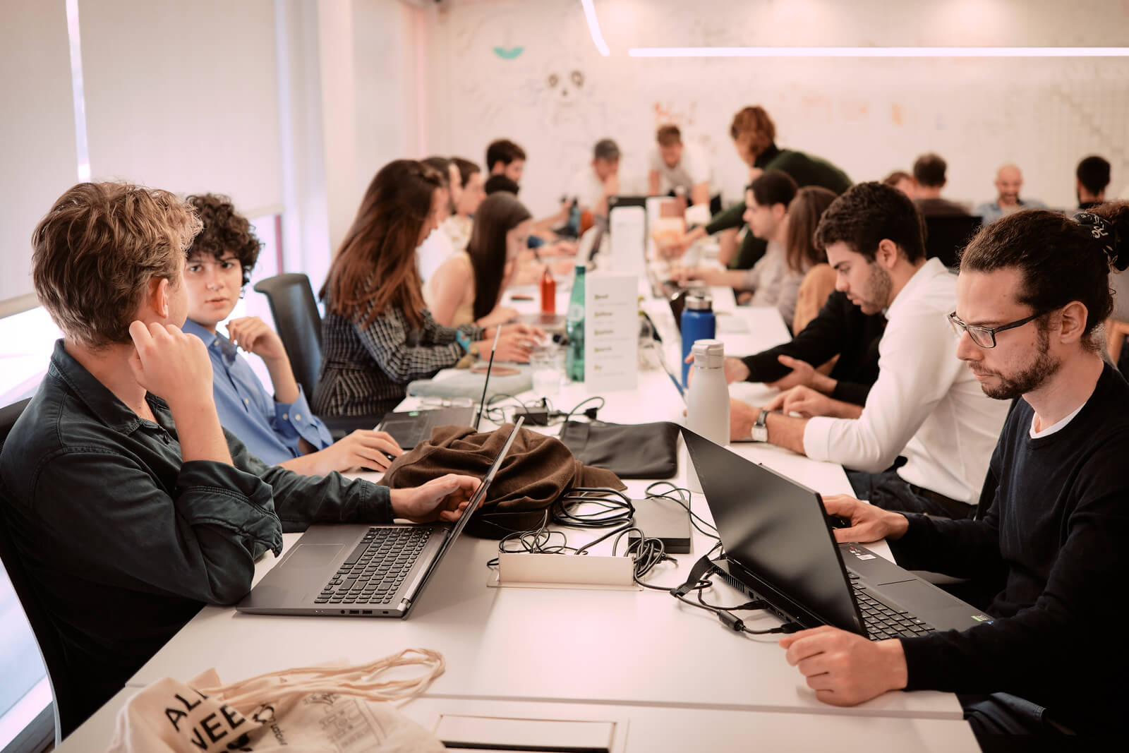 Group of Le Wagon bootcamp participants collaborating and coding on laptops around a shared white table strewn with water bottles, bags and power cables in a modern classroom space