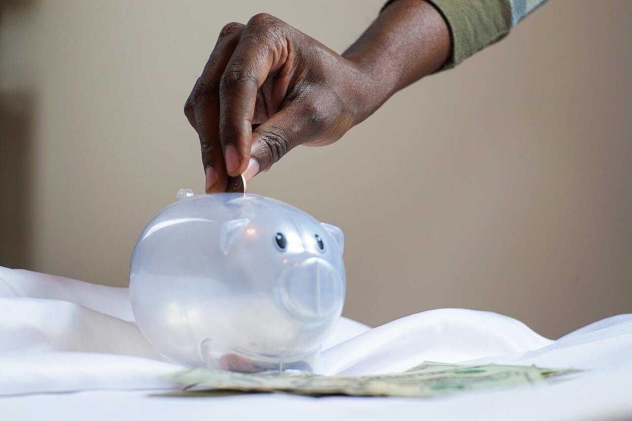 A dark-skinned hand dropping a coin into a clear plastic piggy bank with scattered banknotes on a white cloth background