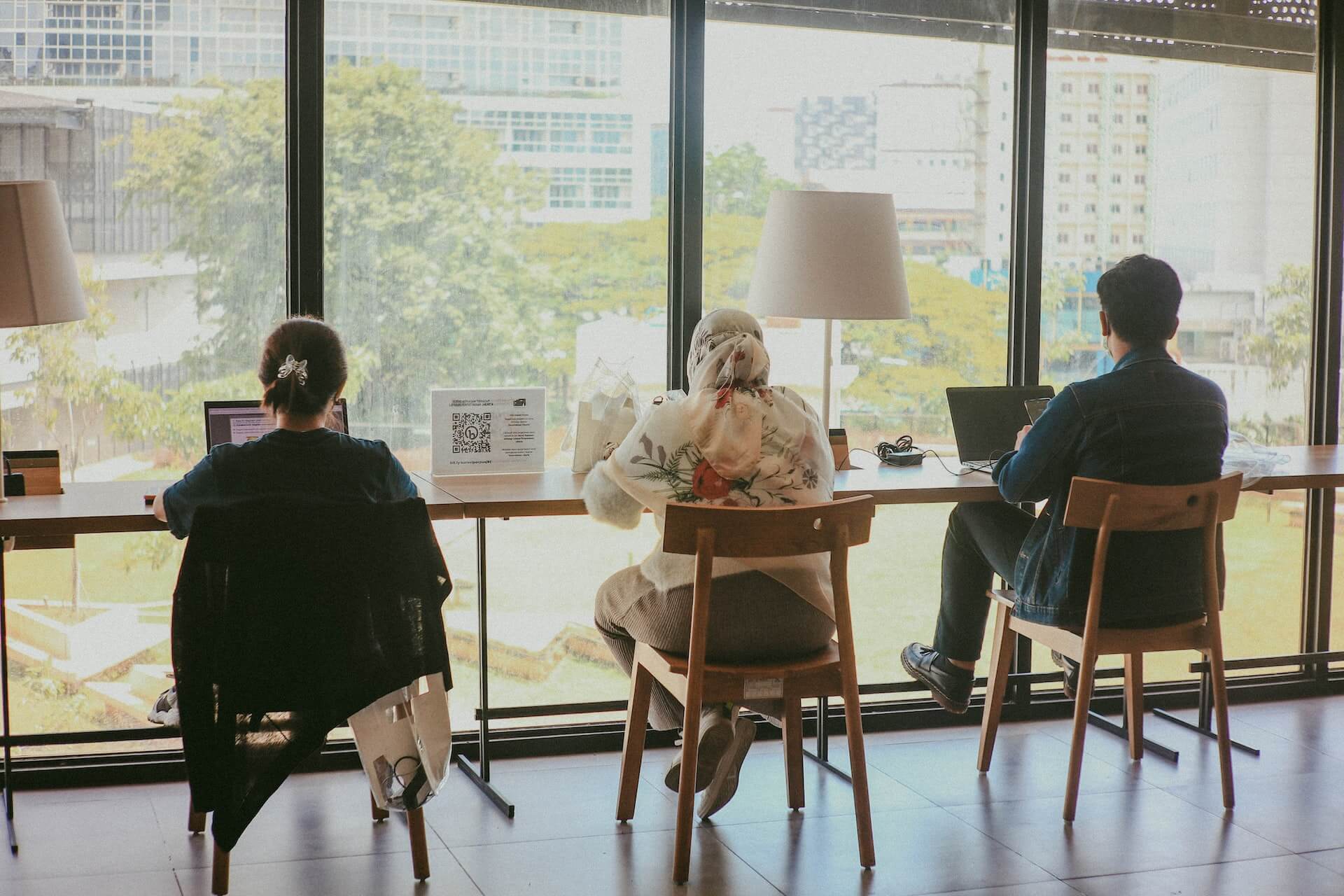 three employees seated at a high wooden desk by a large window using laptops beside modern lamps and a QR code display with an urban park view outside