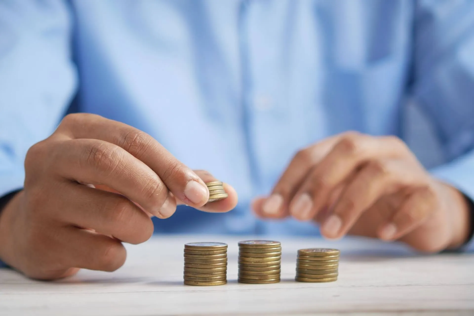 Person in blue shirt arranging stacks of small coins into increasing piles on a wooden table to illustrate saving and budgeting