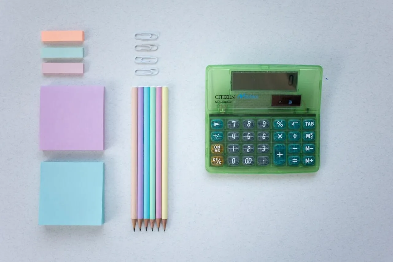 Flat lay of pastel stationery including sticky notes pencils paperclips and a green calculator on a grey desk