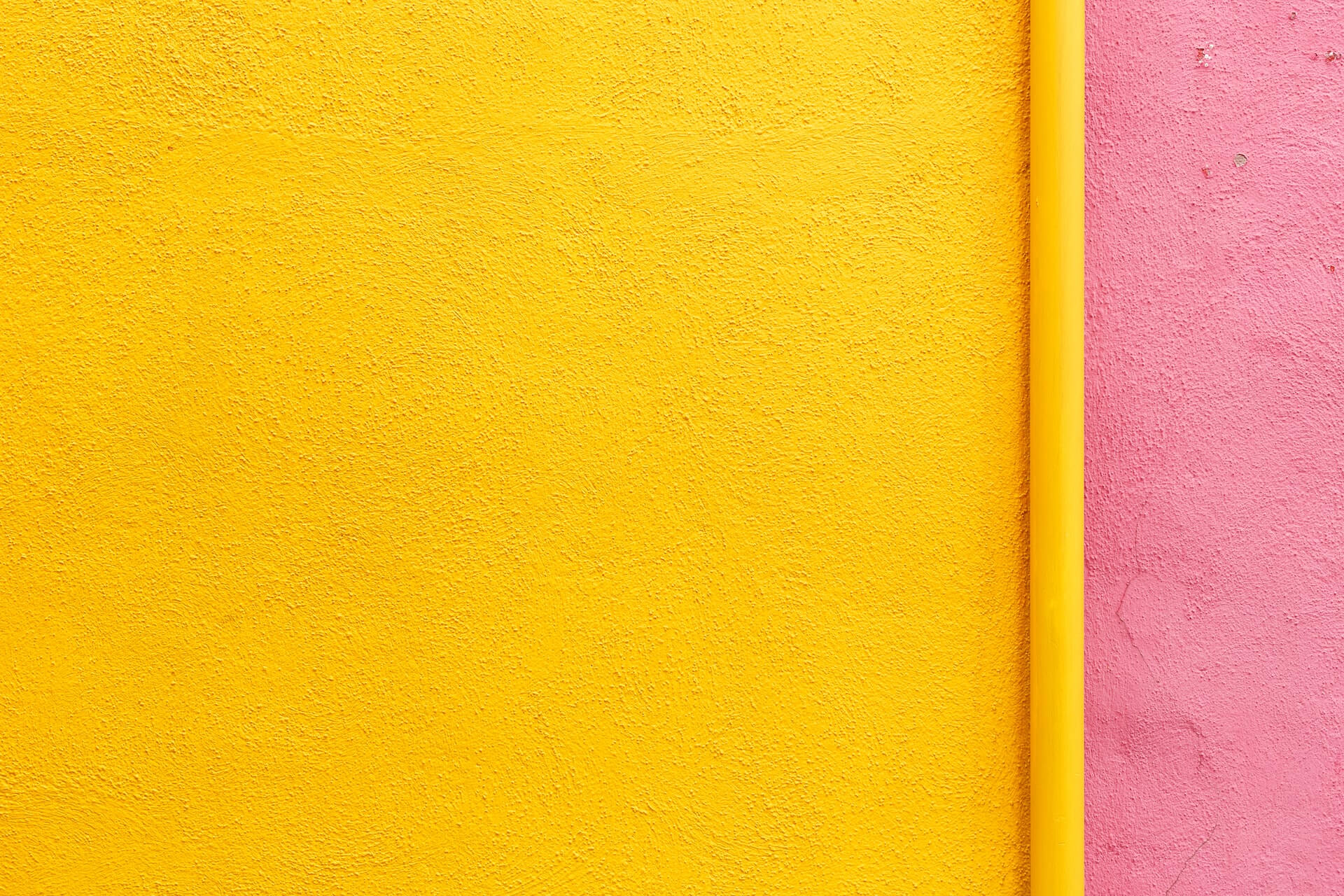 Close-up of a textured yellow wall beside a pink wall with a vertical yellow pipe dividing the two surfaces