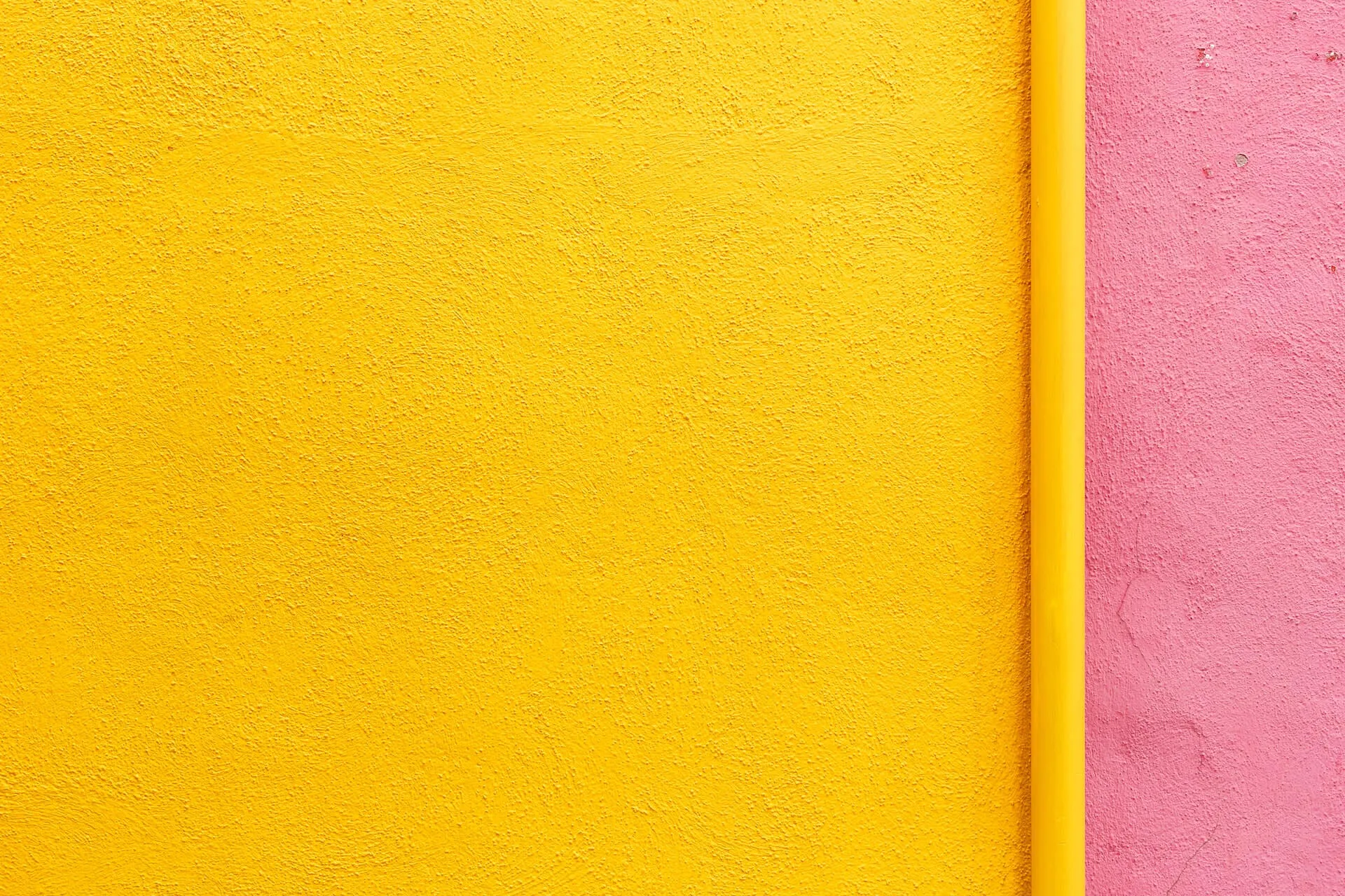 Close-up of a textured yellow wall beside a pink wall with a vertical yellow pipe dividing the two surfaces