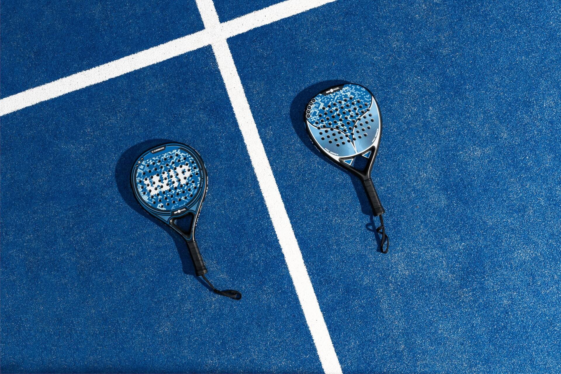 close-up overhead shot of two carbon padel rackets with blue patterned faces and black handles on a blue artificial turf court marked by white boundary lines
