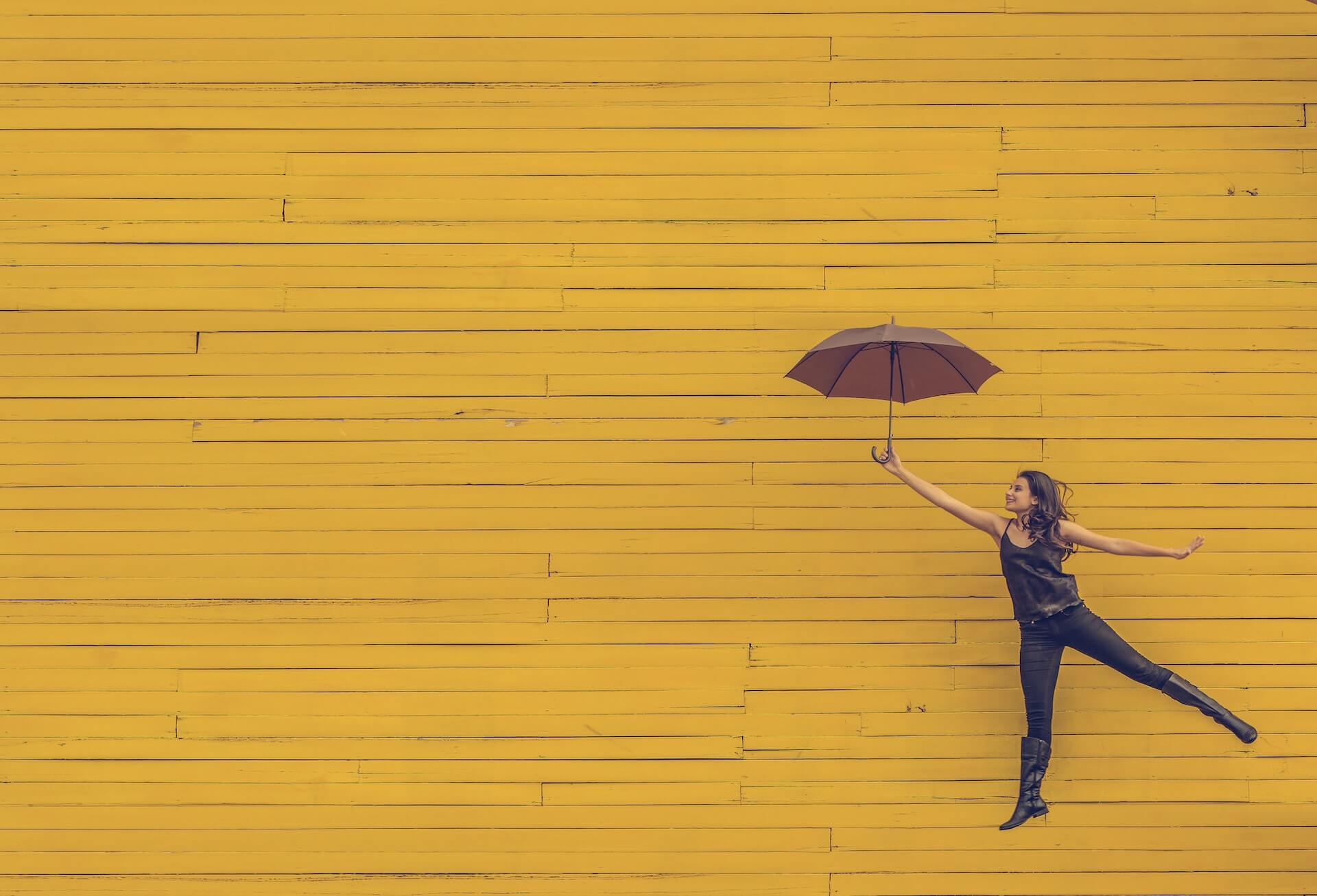 Young woman in black tank top jeans and boots leaps sideways holding a brown umbrella in front of a bright yellow paneled wall evoking playful floating motion