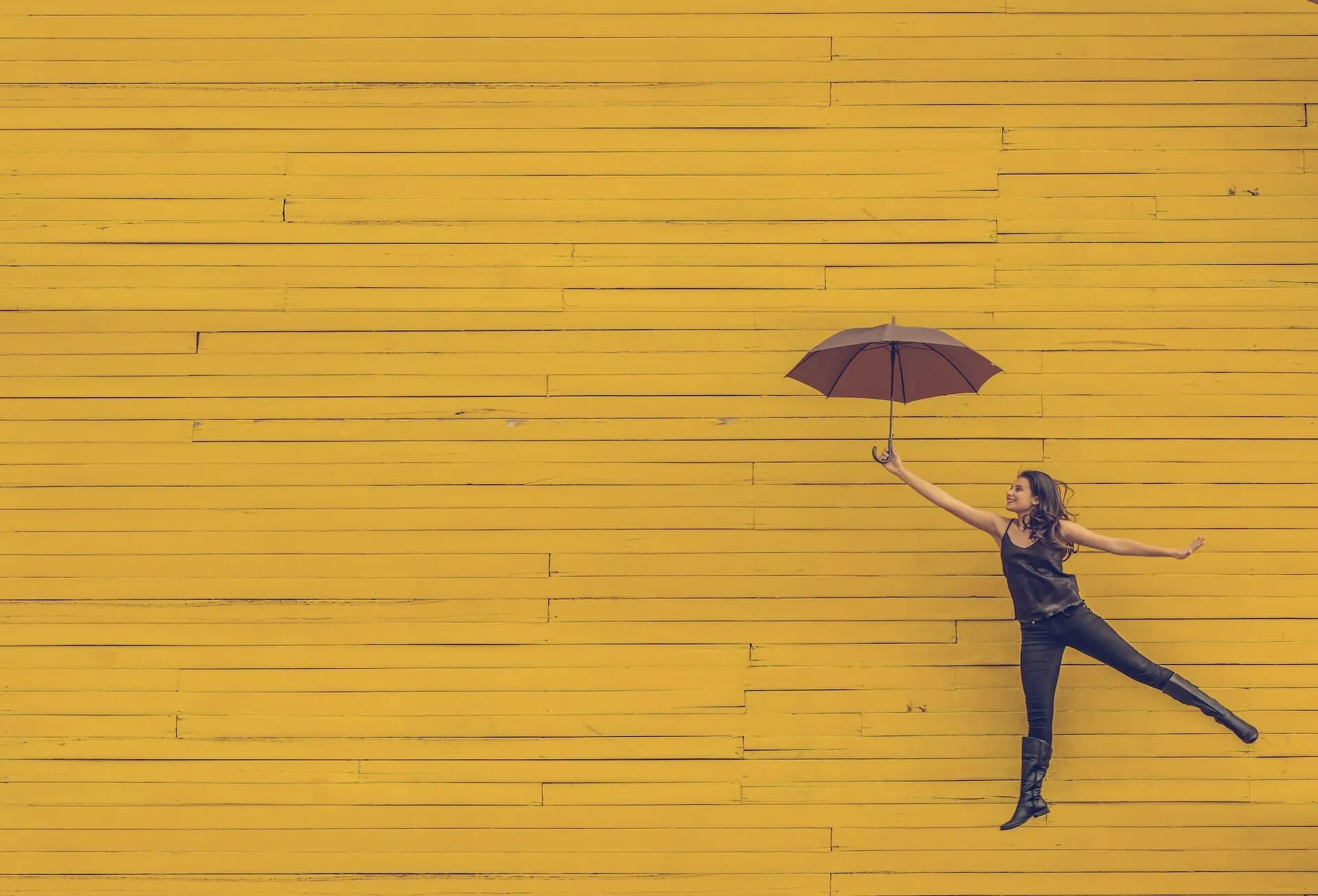 Young woman in black tank top jeans and boots leaps sideways holding a brown umbrella in front of a bright yellow paneled wall evoking playful floating motion