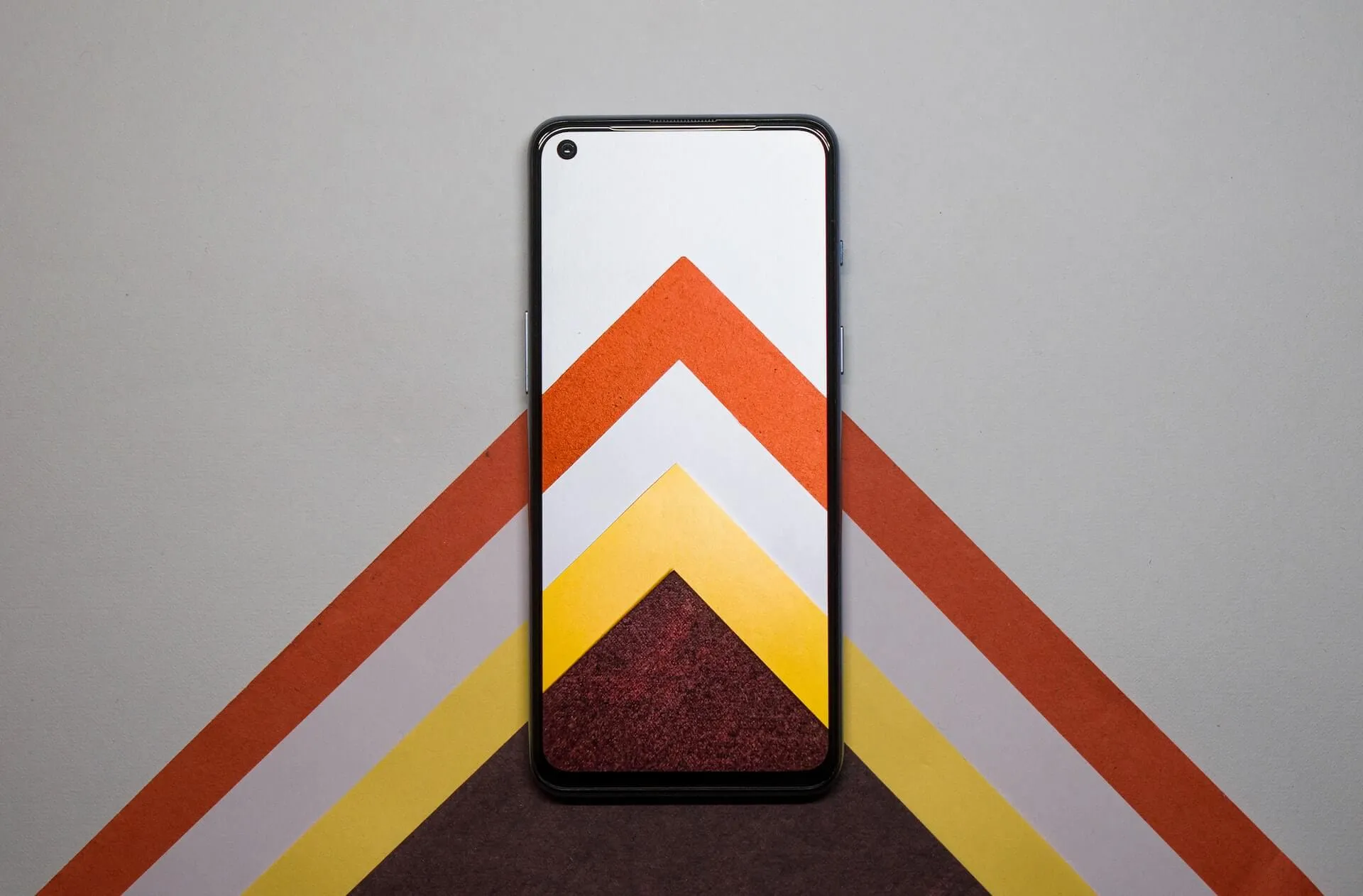 Overhead view of a smartphone centered on a grey background with layered paper chevrons in red, white, yellow and maroon that align with the device screen pattern
