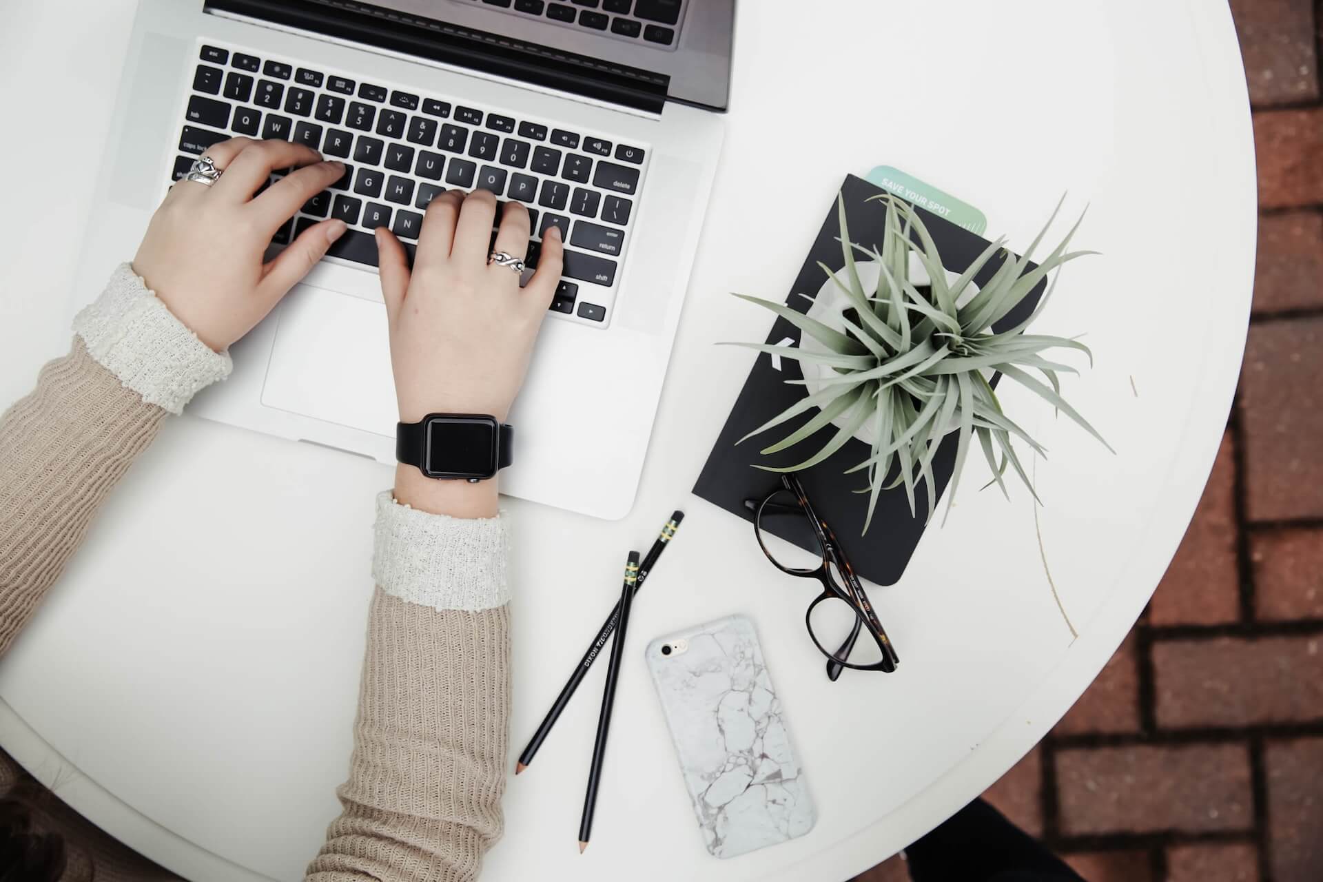 Top-down view of hands wearing a smartwatch typing on a silver laptop at a round white table surrounded by a succulent plant, black notebook with green card, two pencils, eyeglasses and a marble-patterned smartphone