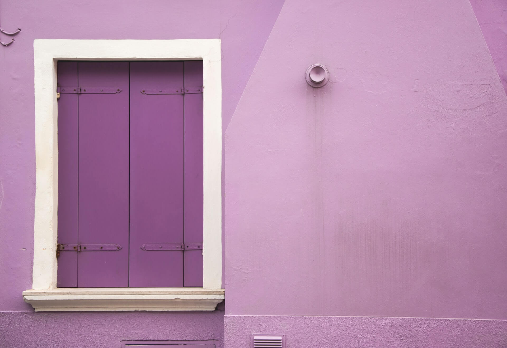 Closed purple wooden shutters within white trim on a pastel lavender facade featuring a small round vent pipe and square wall grate
