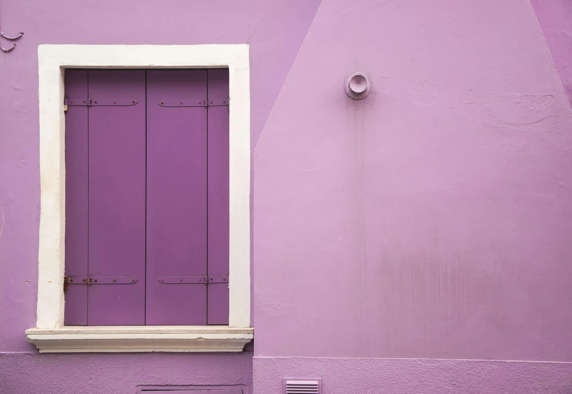 Closed purple wooden shutters within white trim on a pastel lavender facade featuring a small round vent pipe and square wall grate