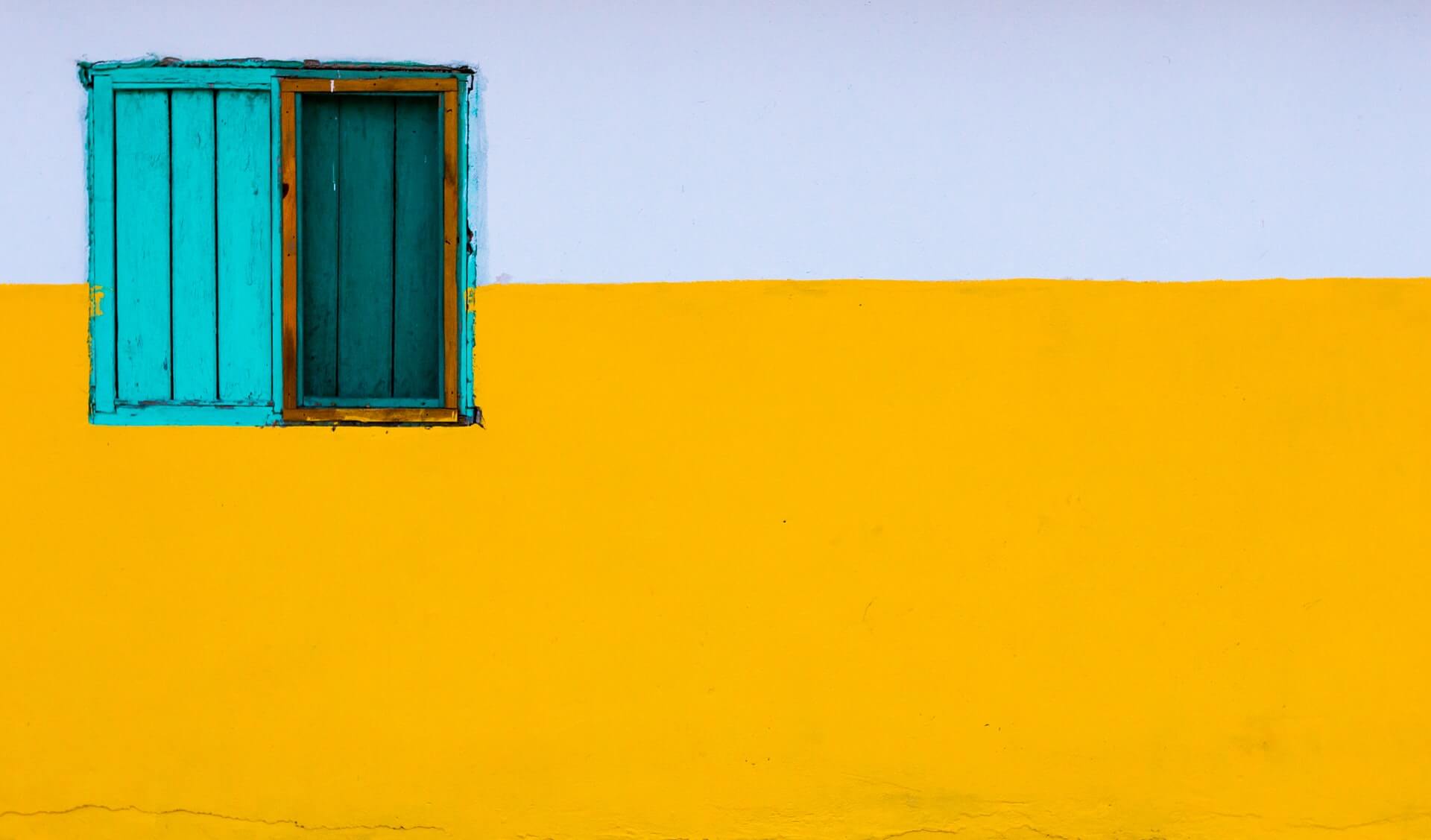 exterior wall painted white on top and yellow on bottom with a turquoise wooden shutter window on the left