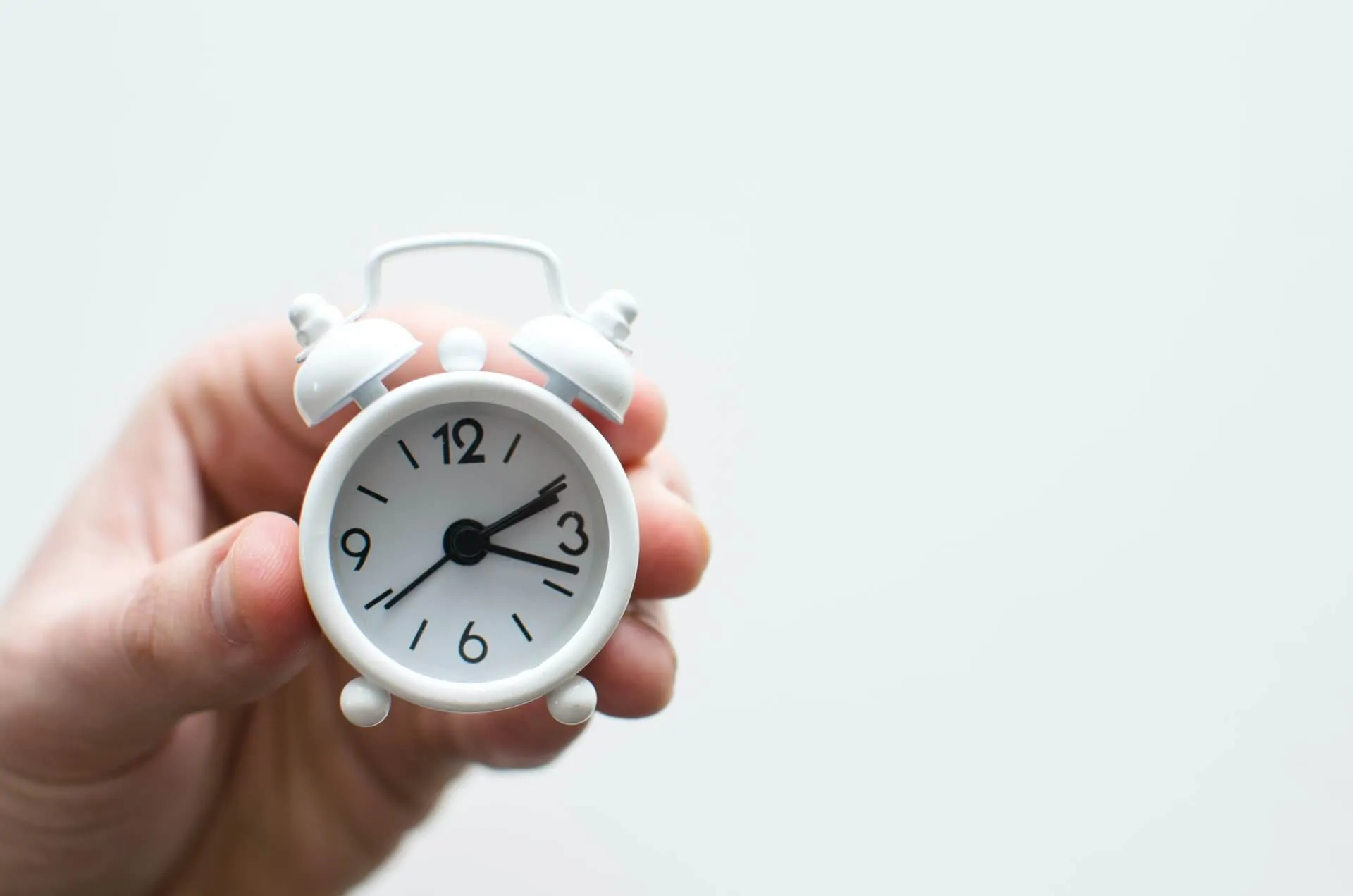 close up of a hand holding a small white analog alarm clock on a plain background suggesting time management